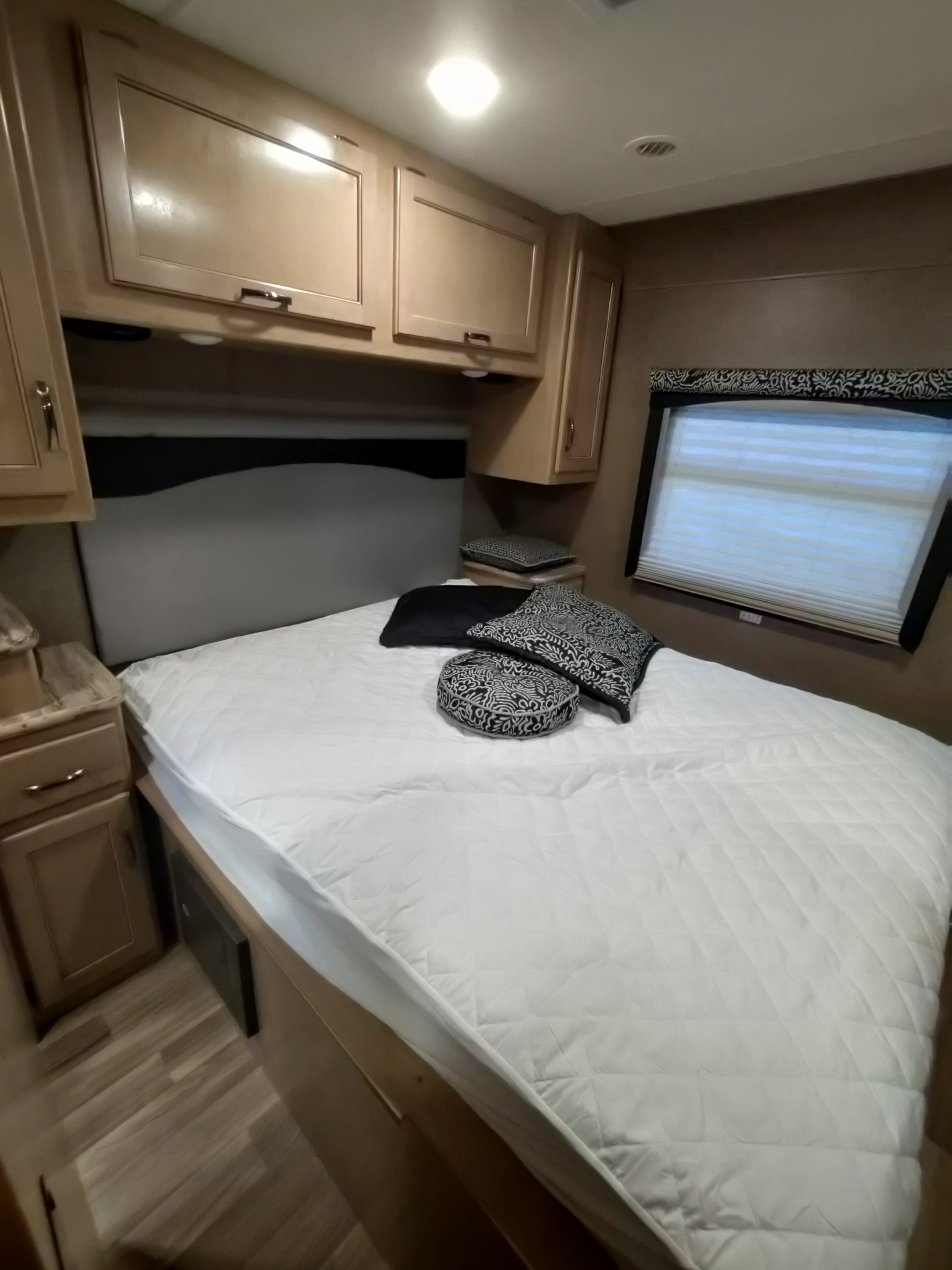 RV rental bedroom showcasing a bed with a white quilted mattress pad, overhead cabinetry, and a window with blinds.. Thor Motor Coach A.C.E 2018