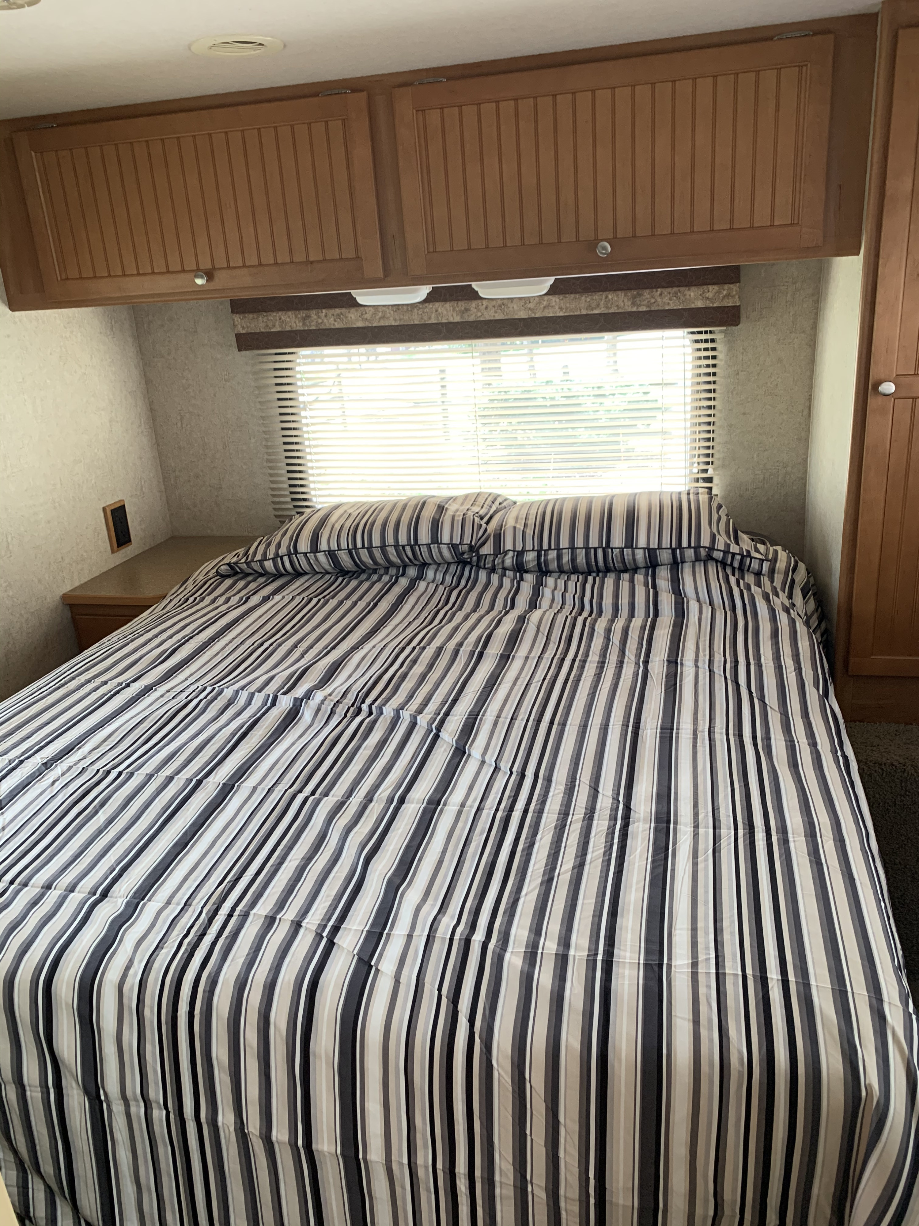 RV rental bedroom featuring a bed with striped bedding, overhead storage, and a window with blinds.. Winnebago Access 2007