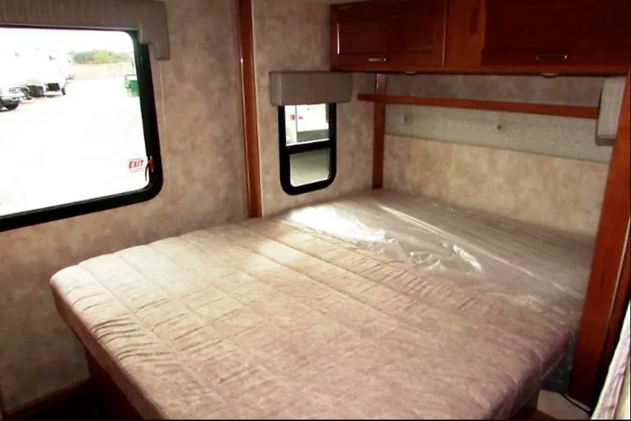 Inside an RV rental, a neat bedroom features a bed with plastic covering, windows, and overhead cabinets.. Winnebago Minnie Winnie 2021