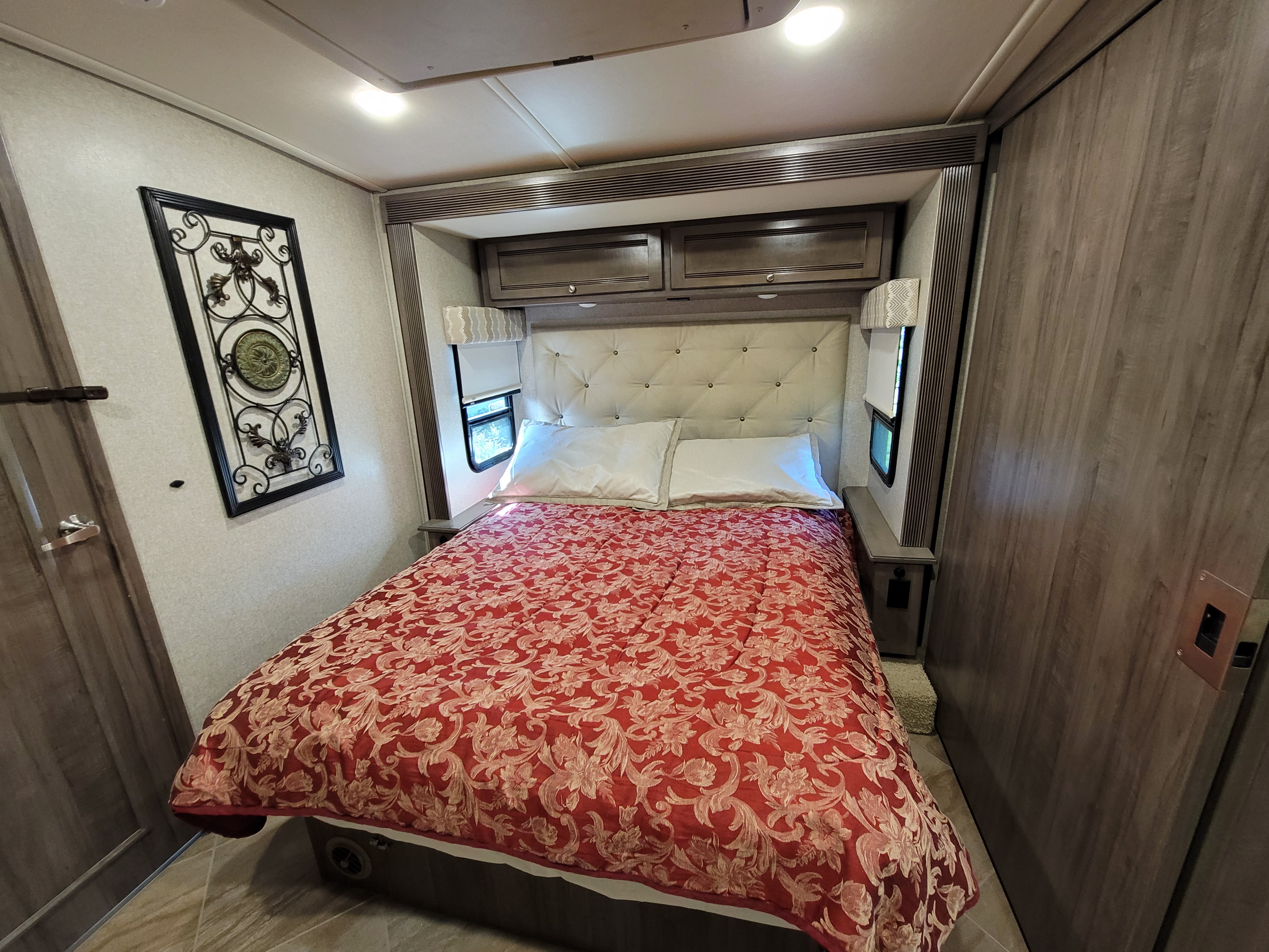 This cozy RV rental bedroom features a bed with a red patterned comforter, tufted headboard, and overhead storage.. Winnebago Forza 2019