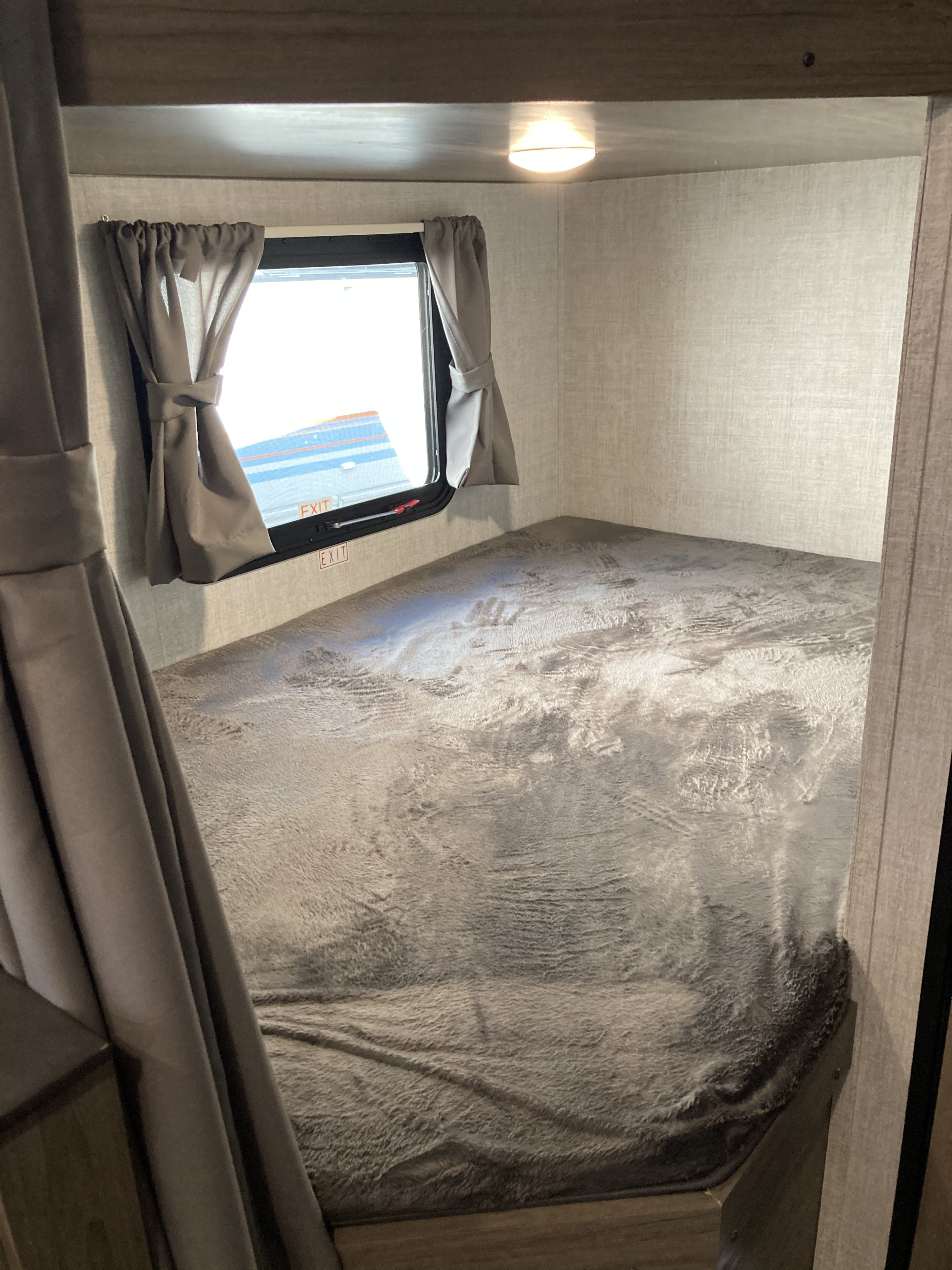 This RV rental features a cozy bunk bed with a textured gray blanket, a small window with tied curtains, and an overhead light.. Gulf Stream Conquest Lite 2021