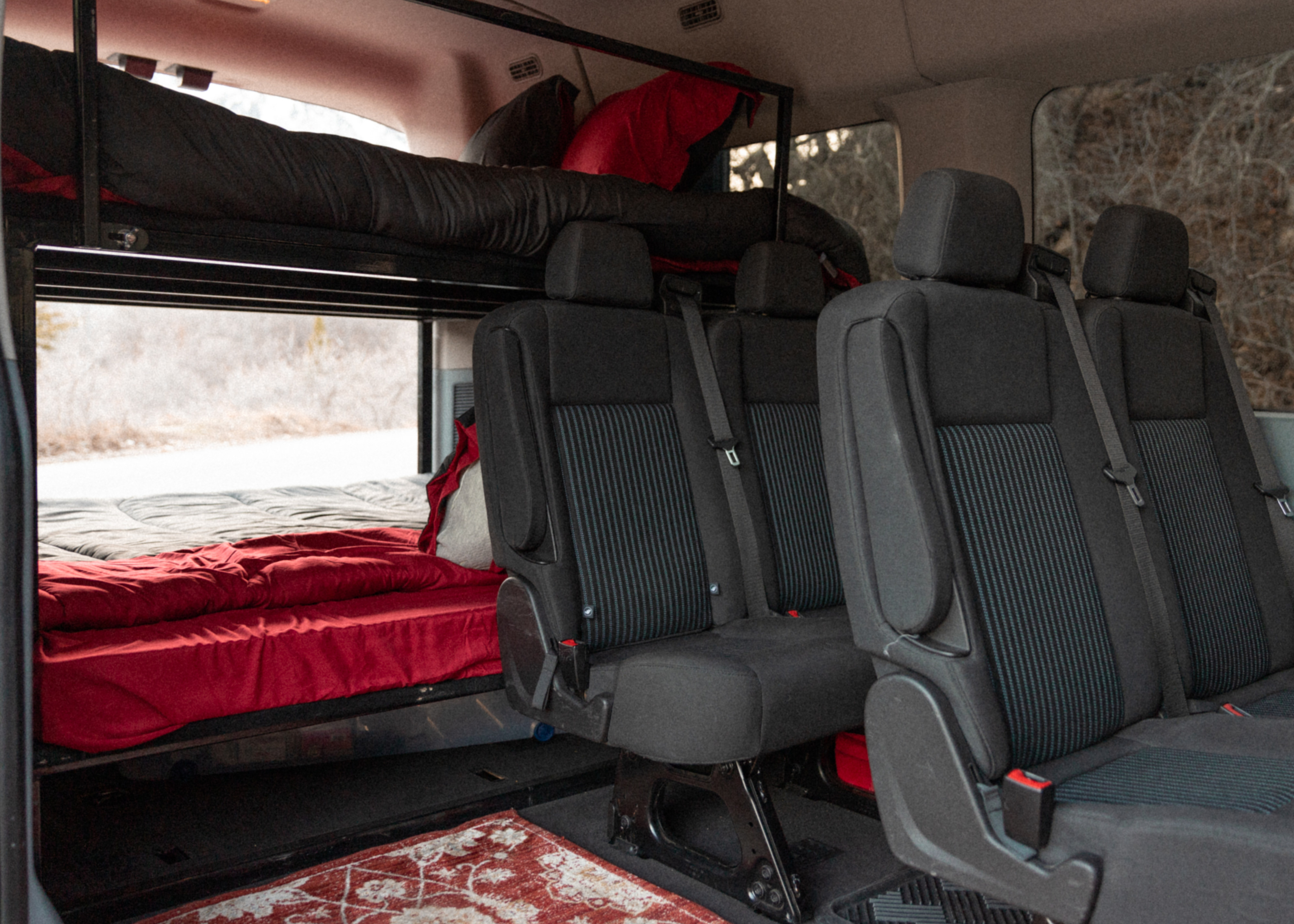 Inside an RV rental van: bunk beds with red and black bedding, rear passenger seats, and an outdoor view, ready for travel.. Ford Transit 2016