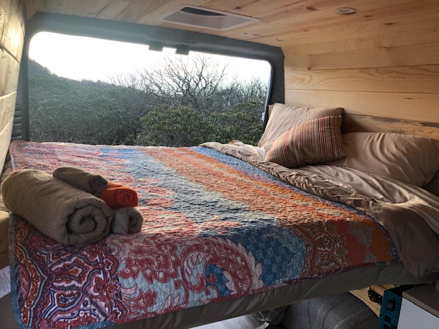 Comfortable rv rental bed with colorful quilt, rolled towels, and a beautiful natural view from the large window.. Ram Promaster 2021