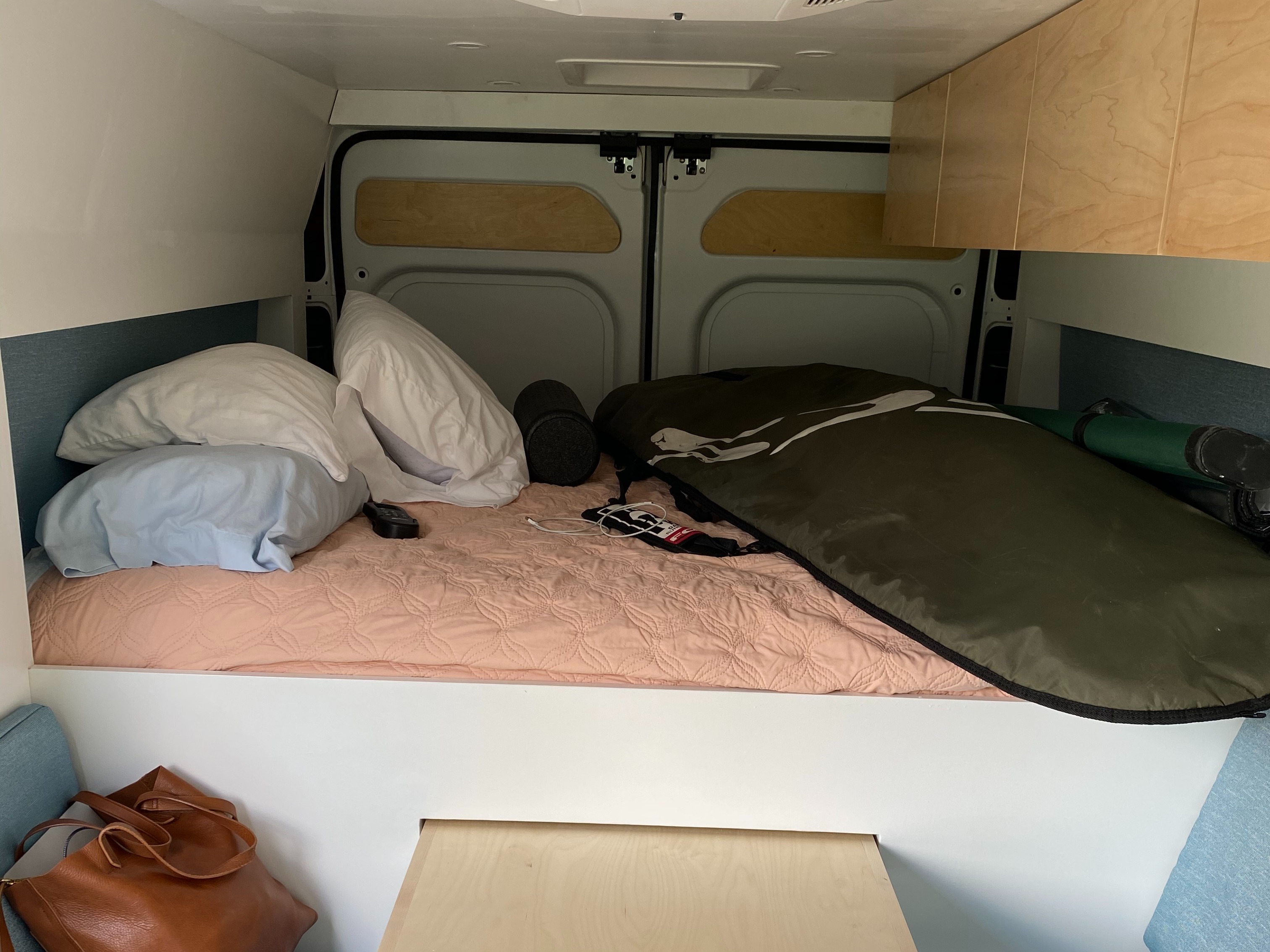 Cozy bed setup in an rv rental, featuring pillows, a surfboard bag, and overhead cabinets for storage in a converted van.. Dodge Other 2020