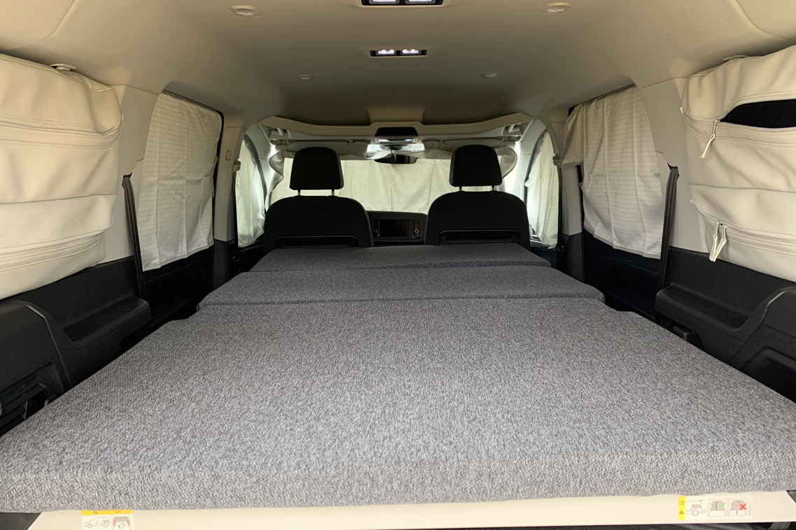 View inside an RV rental, featuring a large grey bed, side storage, and window curtains for a comfortable mobile living space.. Volkswagen Caddy 2021
