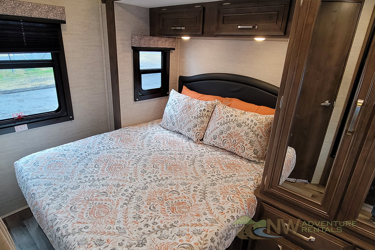 RV rental bedroom featuring a comfy bed with patterned quilt, dark headboard, two windows, and wooden cabinetry.. Jayco Melbourne 2021