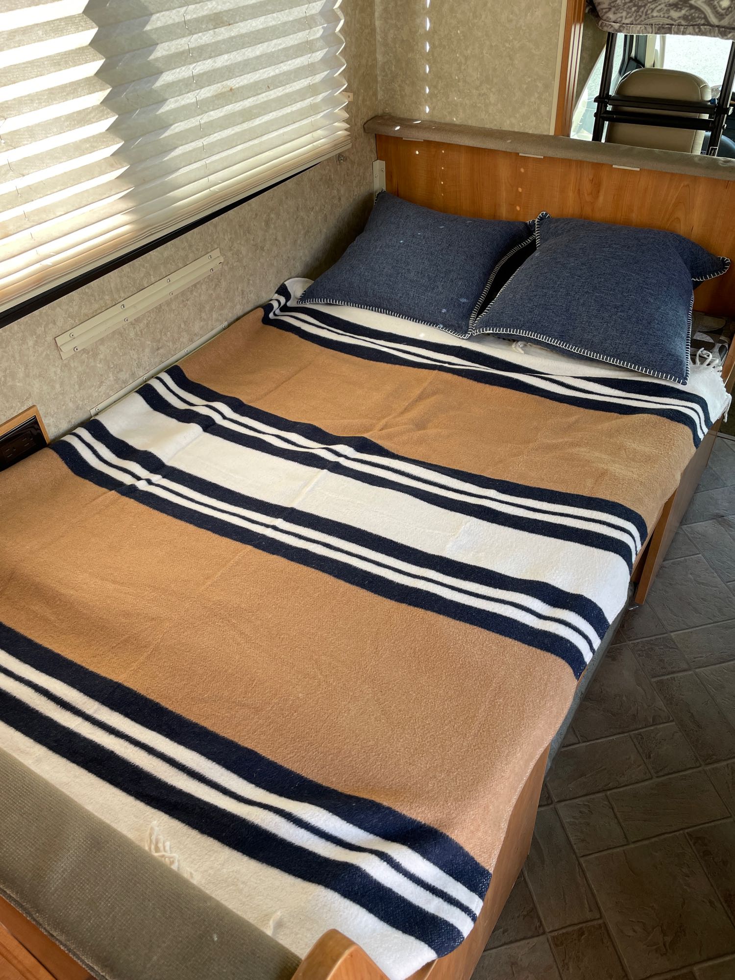 Cozy RV rental sleeping area with a striped blanket, blue pillows, and window with blinds.. Winnebago Access 2010