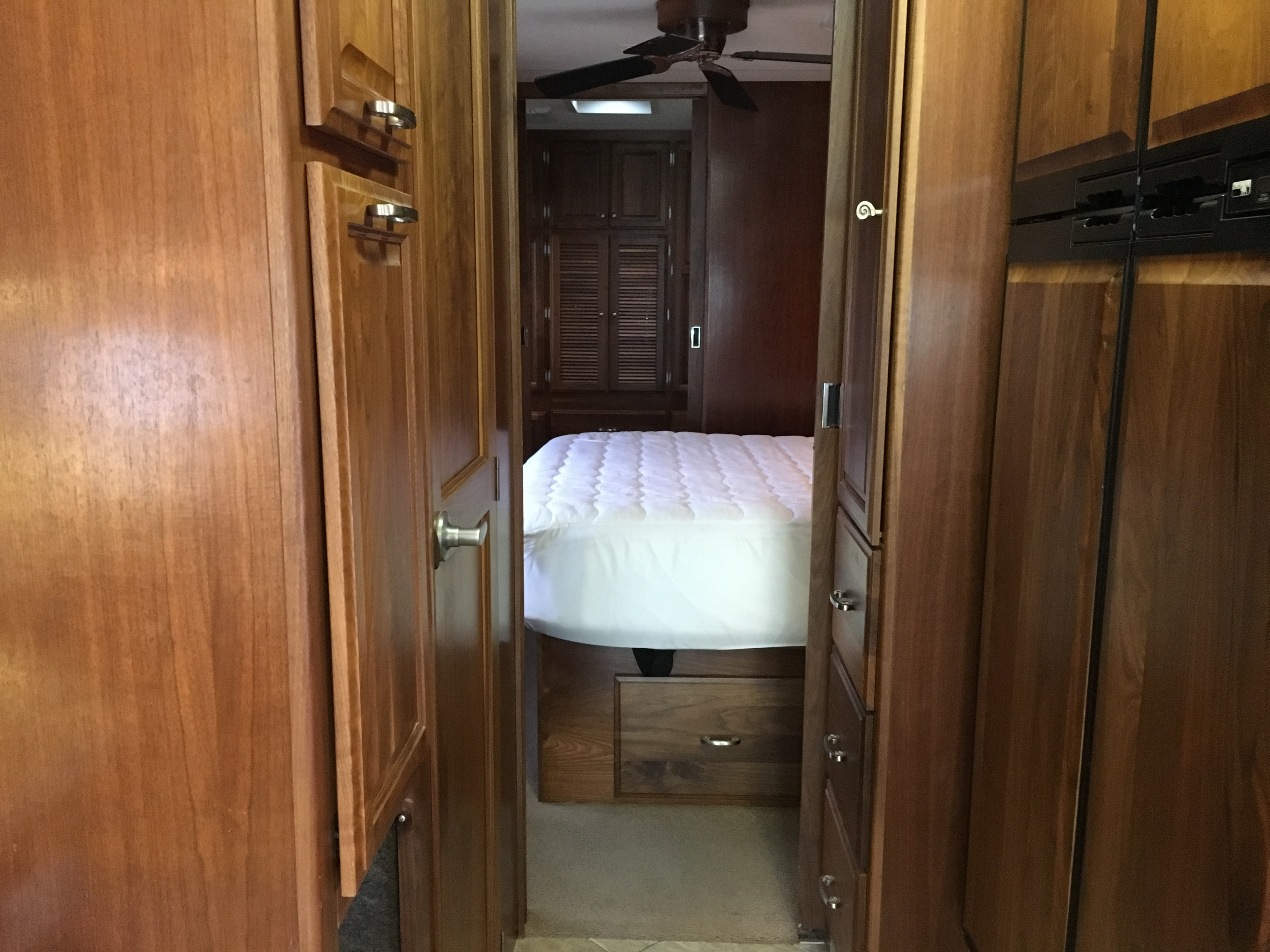 Cozy rv rental interior featuring a wooden hallway leading to a bedroom with a white bed and ceiling fan.. Fleetwood Revolution Le 2005