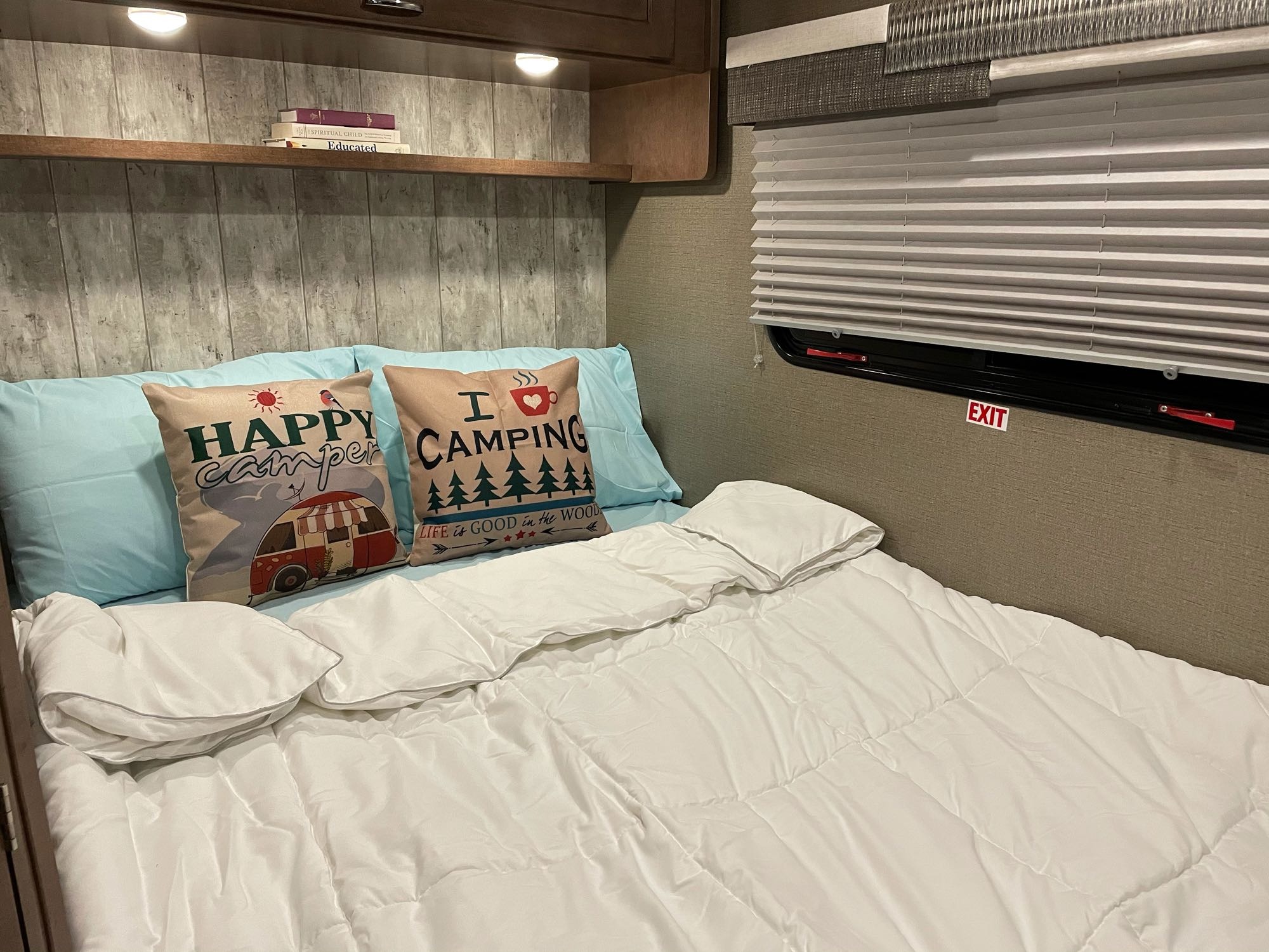 RV rental bedroom features a comfortable bed, 'Happy Camper' pillows, and a shelf with books, complete with an "EXIT" sign.. Winnebago Outlook 2019