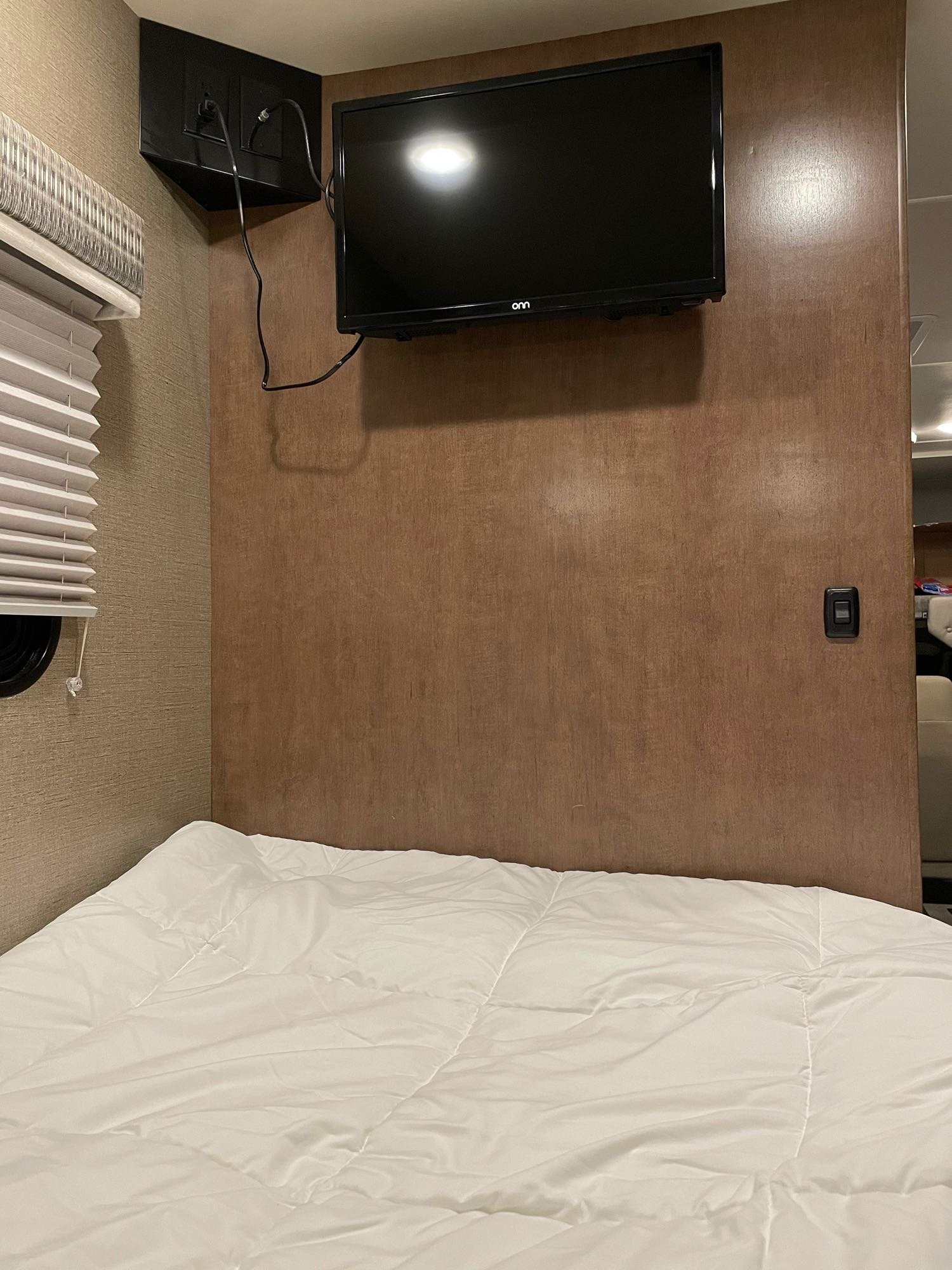 RV rental bedroom features a wall-mounted TV over a white bed with window blinds. Compact and functional space.. Winnebago Outlook 2019