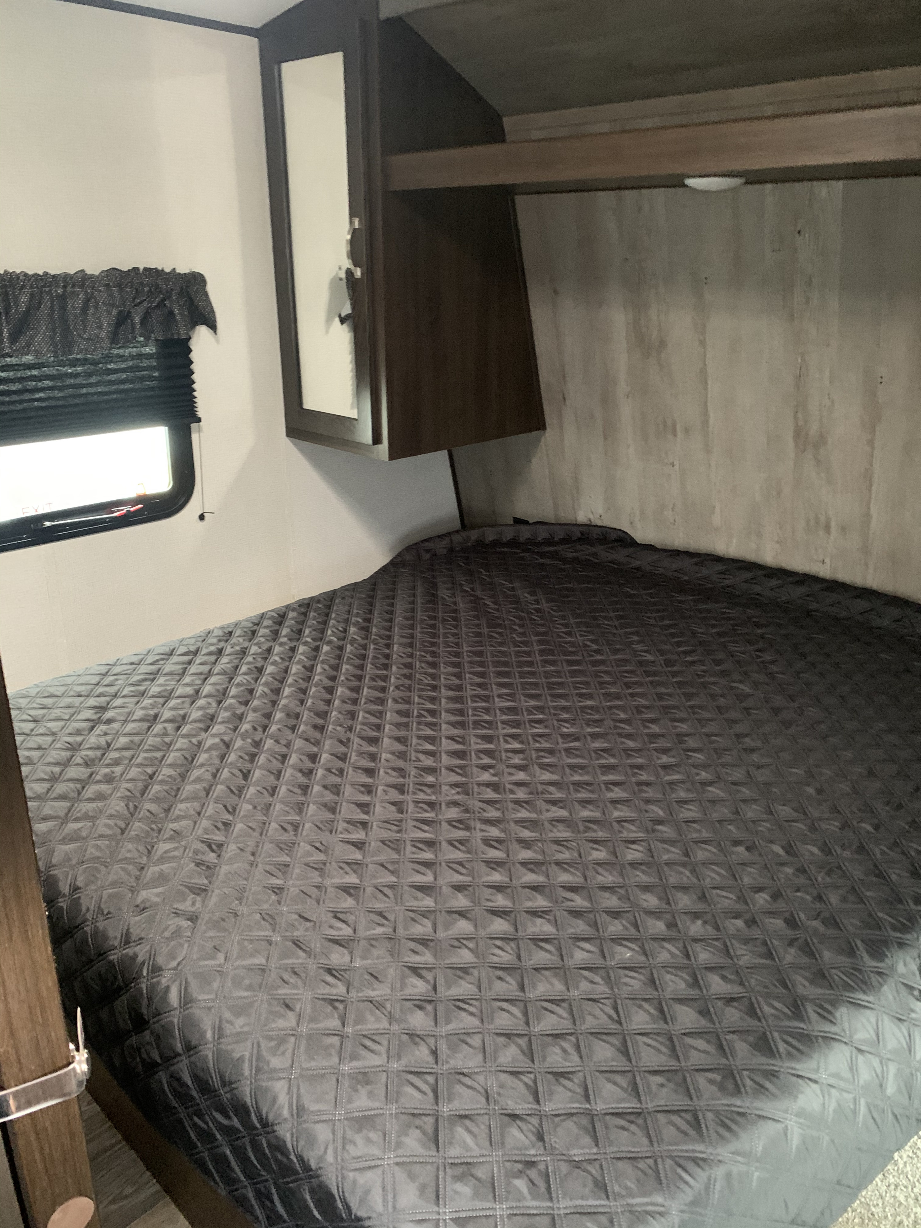 Inside an RV rental, a compact bedroom features a large, dark gray quilted bed, overhead cabinet, and window.. Keystone Springdale 2021