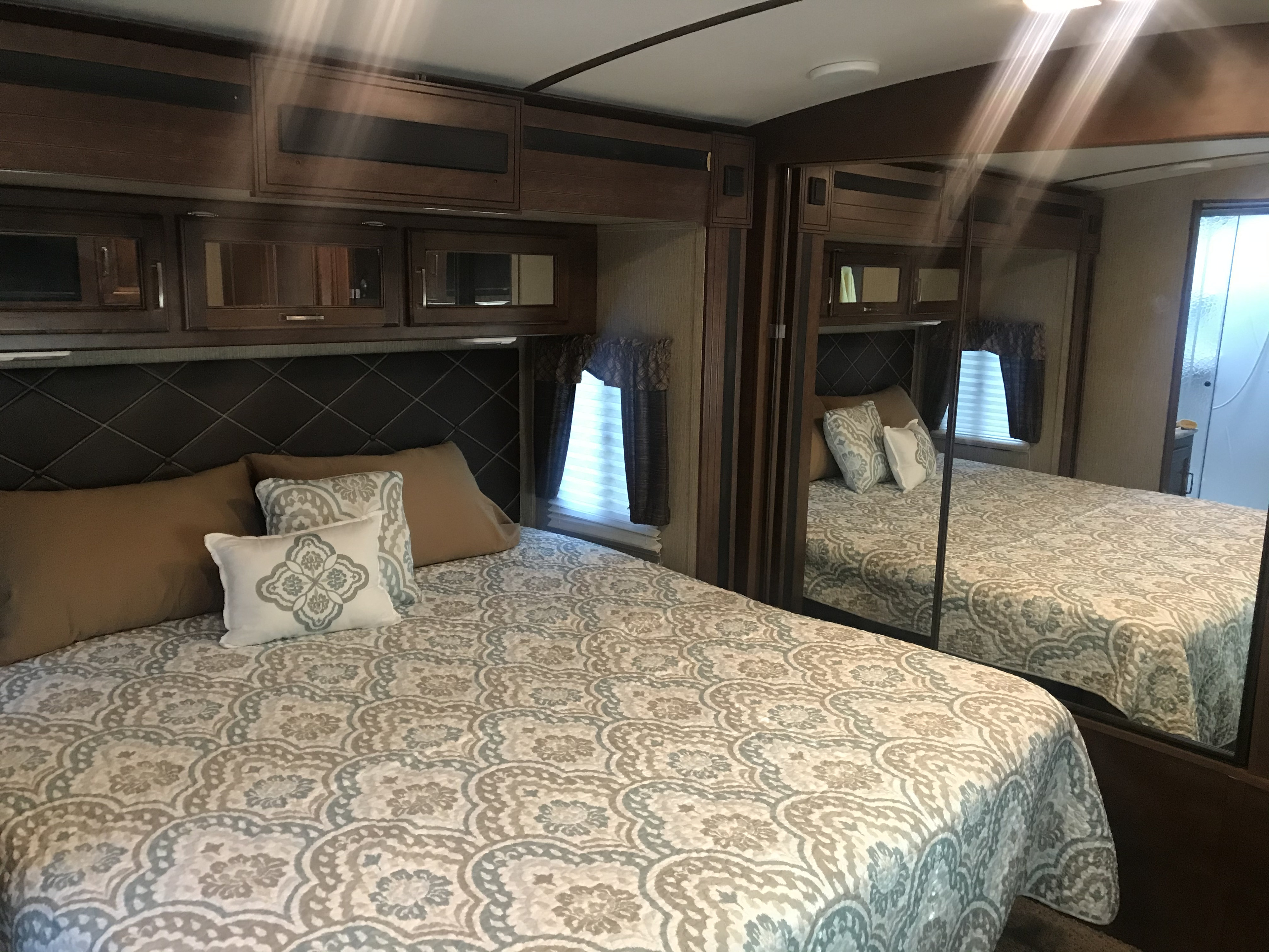 Cozy RV rental bedroom featuring a bed with elegant patterned bedding, dark wood cabinets, and a large mirrored closet.. Keystone Outback 2018
