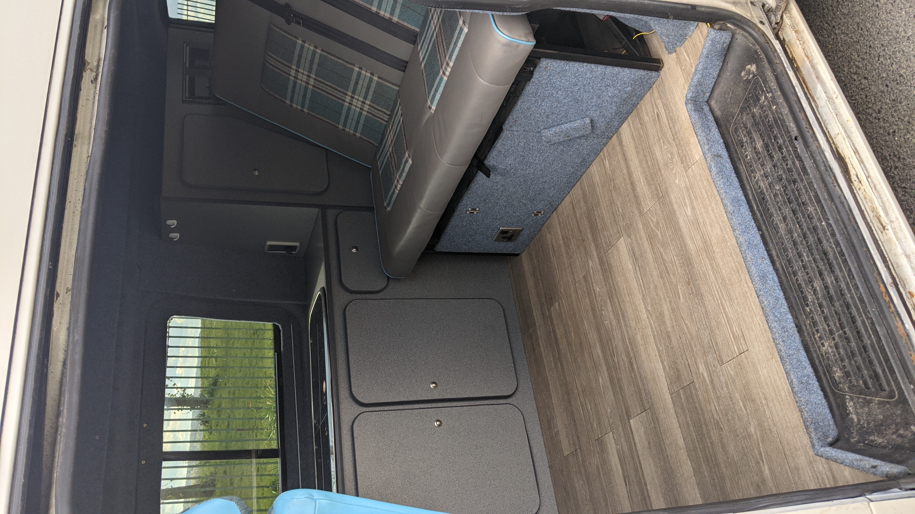 RV rental interior: A grey bench with blue plaid accents, wood-look floor, and built-in storage in a cozy camper van.. vw T5 2004