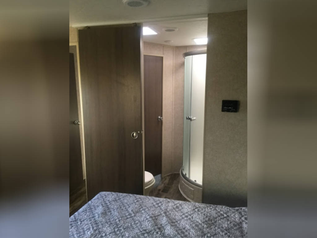 An RV rental interior showing a cozy bedroom with patterned bedding, leading to a compact bathroom featuring a toilet and shower.. Coachmen Catalina 2020