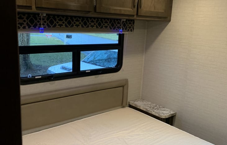 Inside an RV rental bedroom. Features a bed, window with outdoor view, overhead cabinets & small nightstand. Blue lighting detail.. Jayco Redhawk 2020