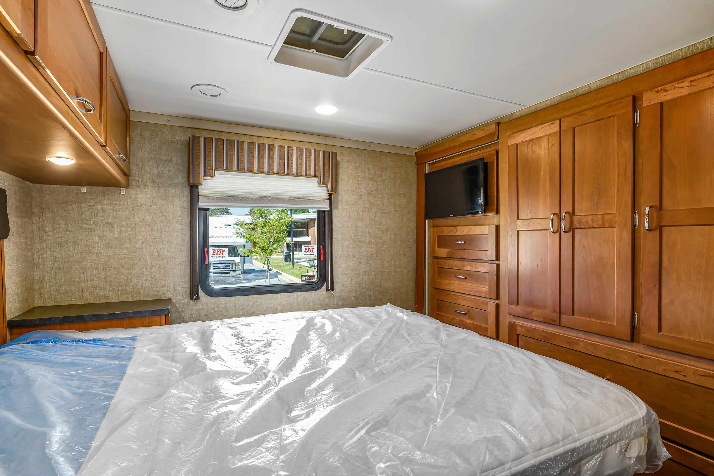 A bright RV rental bedroom features a plastic-wrapped bed, wooden cabinets, a TV, and a window view.. Winnebago Spirit 2017