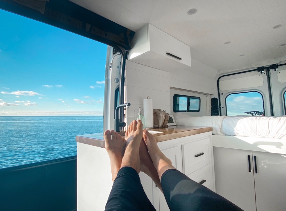Legs up, enjoying the vast ocean view from inside a bright, modern RV rental van. Perfect for remote work or vacation.. Ram Promaster 2500 2016