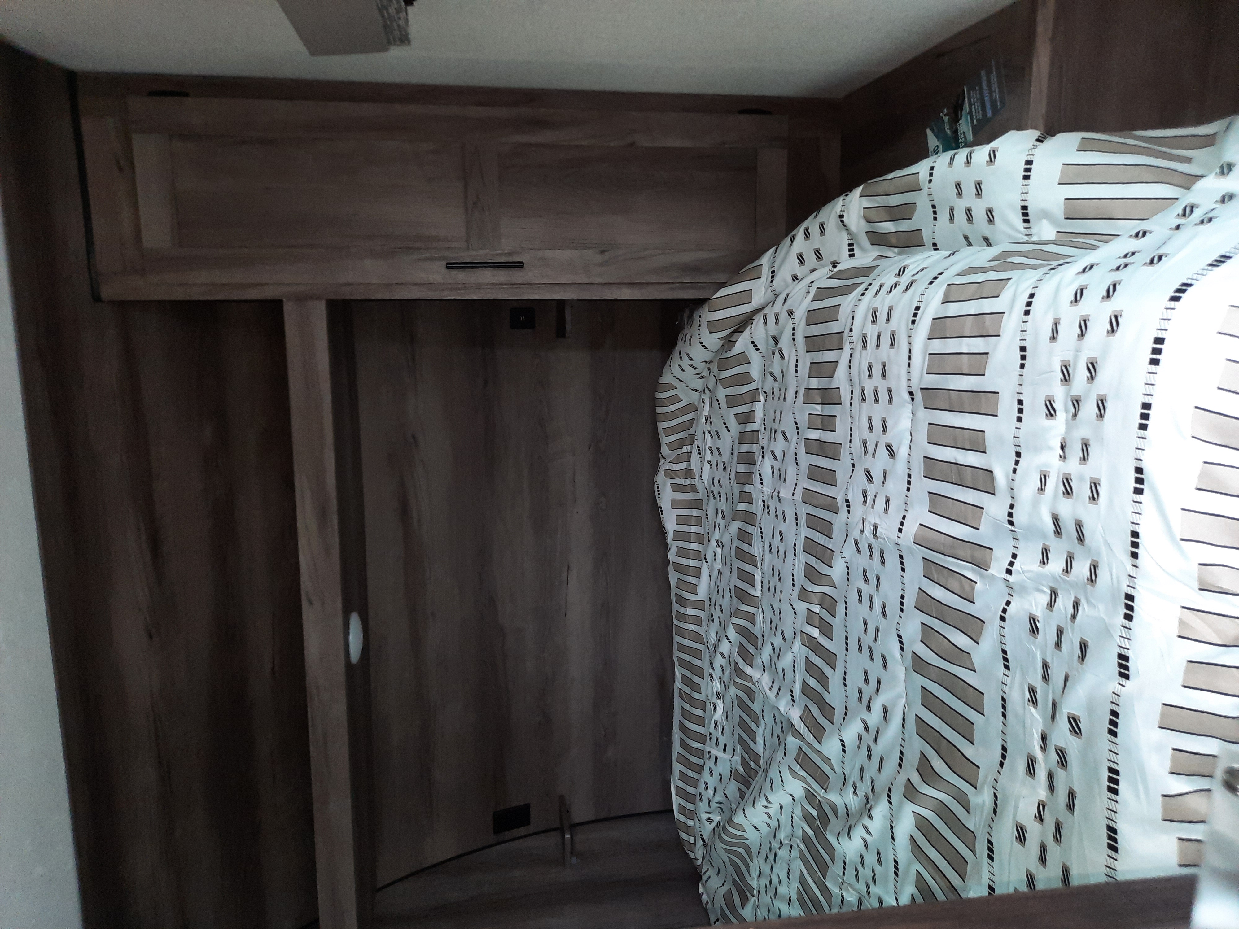 Cozy RV rental bedroom featuring a patterned bed, overhead wooden cabinets, and wall paneling, ideal for travel accommodation.. K-Z Manufacturing Sportsmen 2021