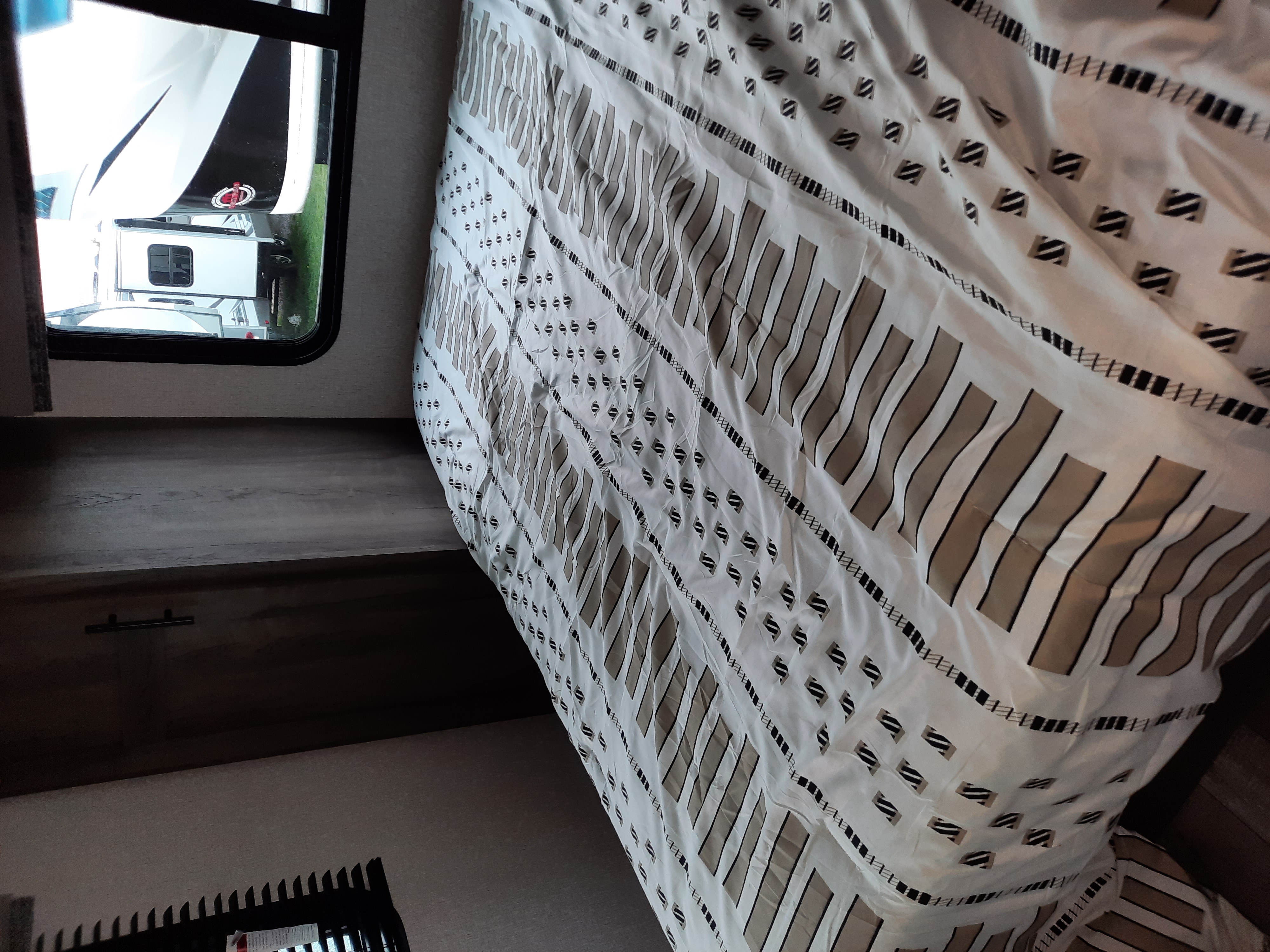 RV rental interior: A white and tan patterned comforter on a cozy bed, with another RV visible through the window.. K-Z Manufacturing Sportsmen 2021