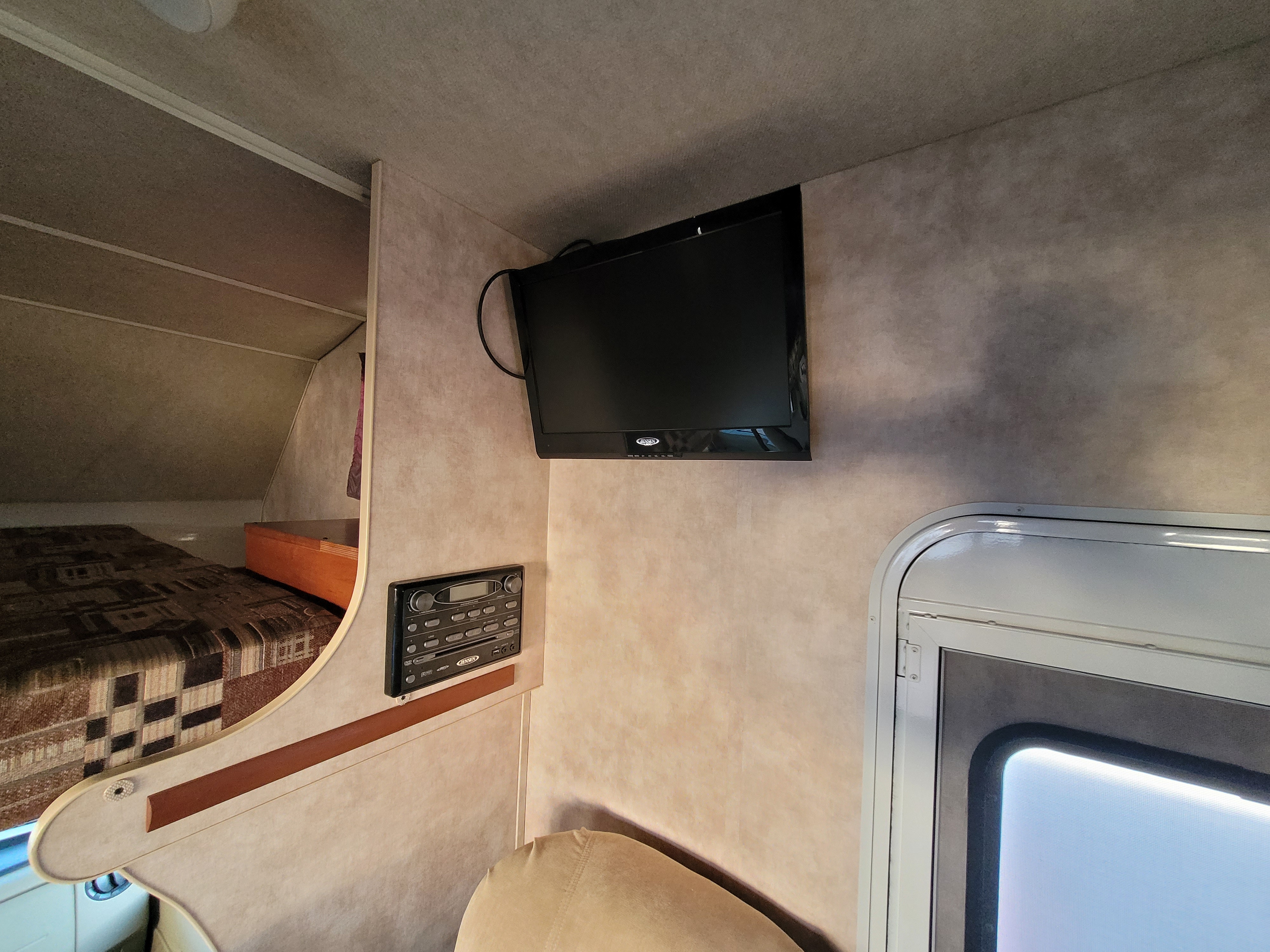 Cozy RV rental interior showcasing a wall-mounted TV, stereo system, elevated sleeping bunk, and a framed window.. Winnebago Chalet 2013