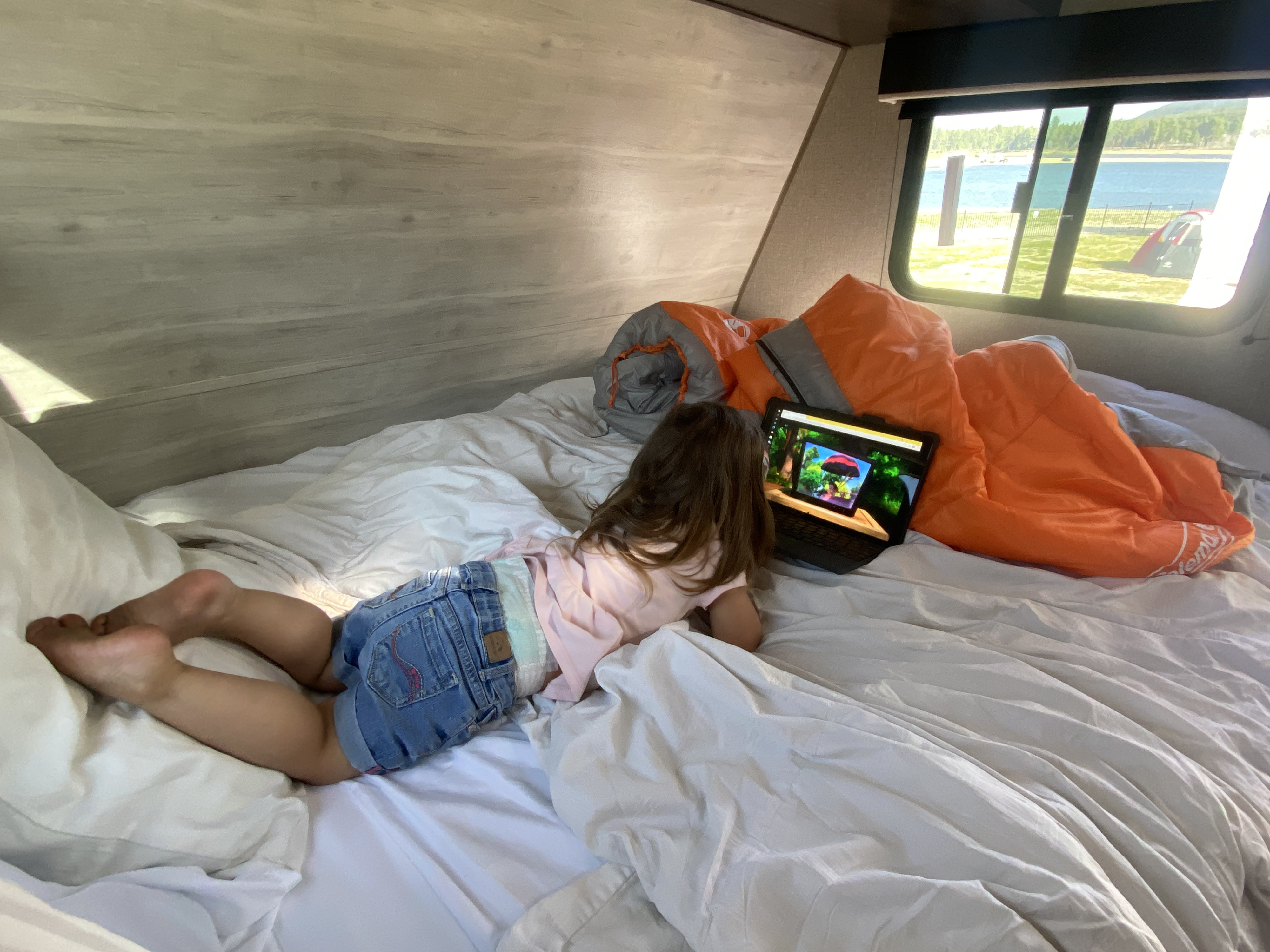 Inside a spacious RV rental, a child lies on the bed watching a tablet, with a serene lake and mountains visible outside.. Jayco Jay Flight 2021