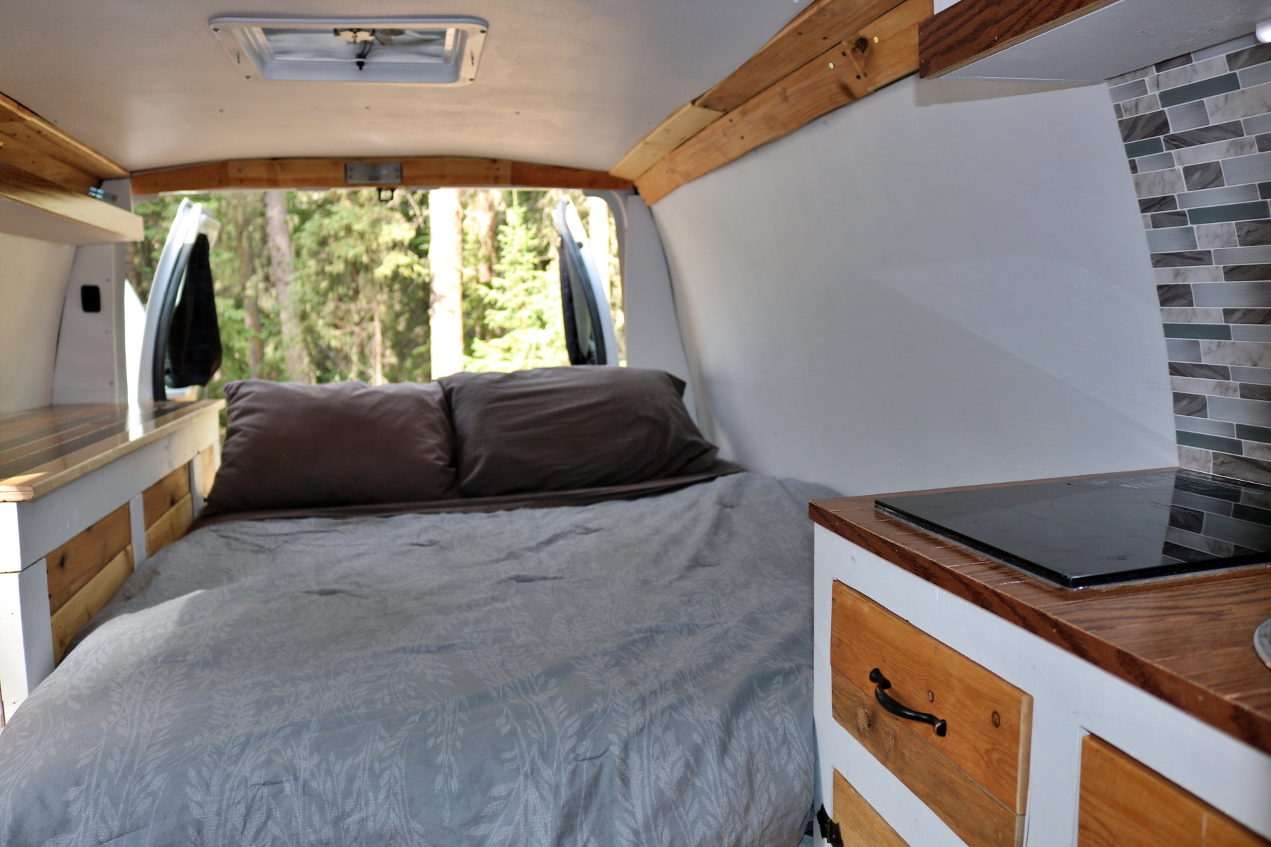 Cozy rv rental interior featuring a comfortable bed, compact kitchen with cooktop, and a scenic forest view from the back.. Ford Econoline 2009