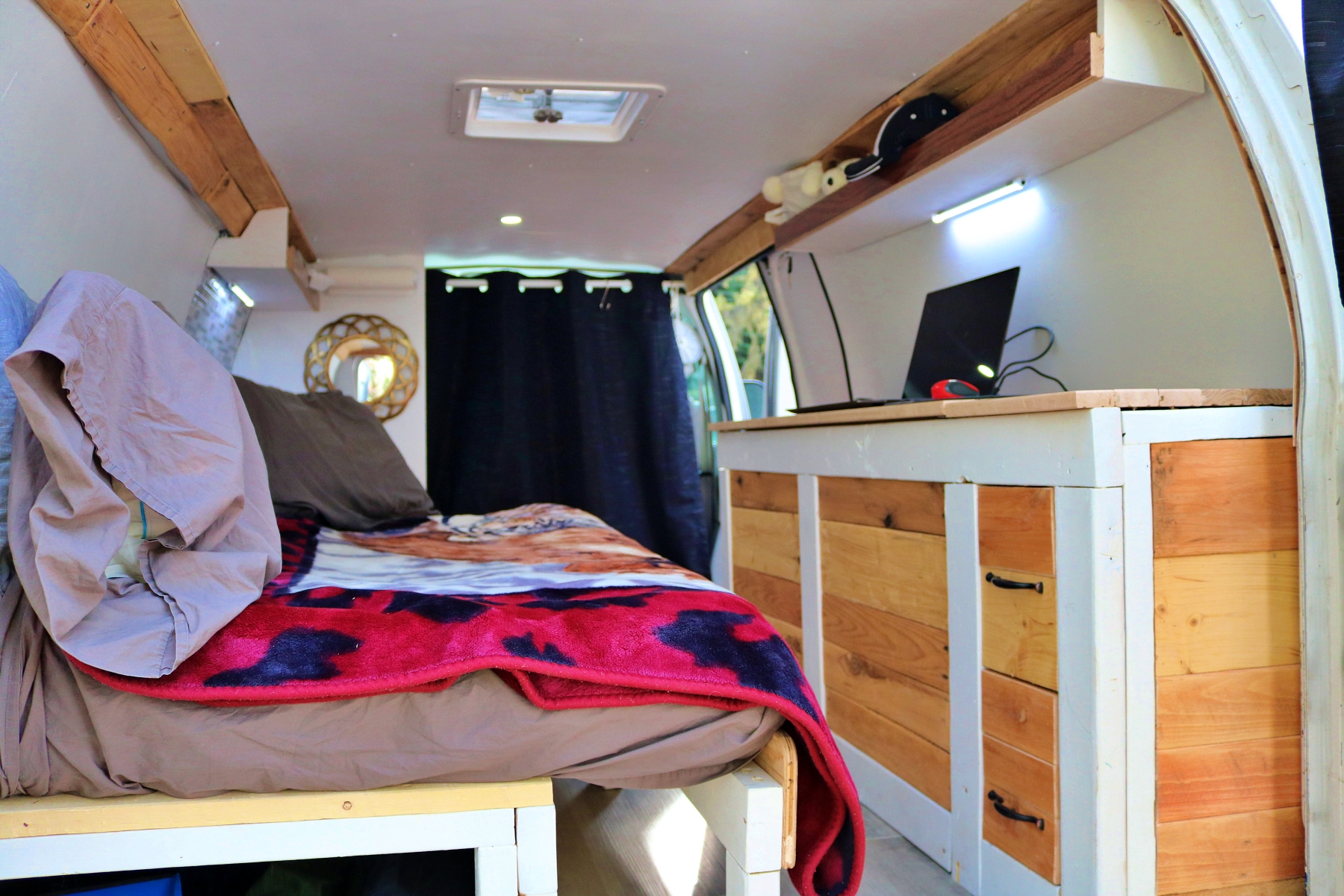 This RV rental features a cozy van interior with a bed, a wooden counter holding a laptop, and overhead shelving.. Ford Econoline 2009