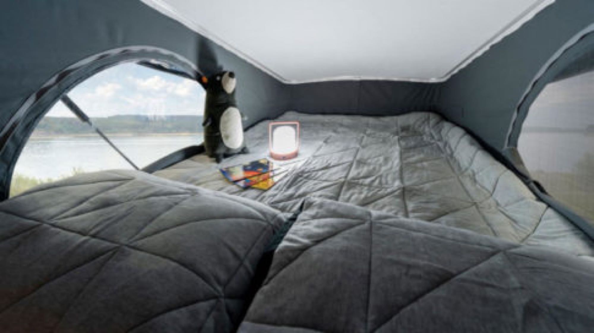 Inside a cozy RV rental pop-top, a gray bed faces a scenic lake view window. Features a stuffed animal & lit lantern.. Winnebago Solis 2021