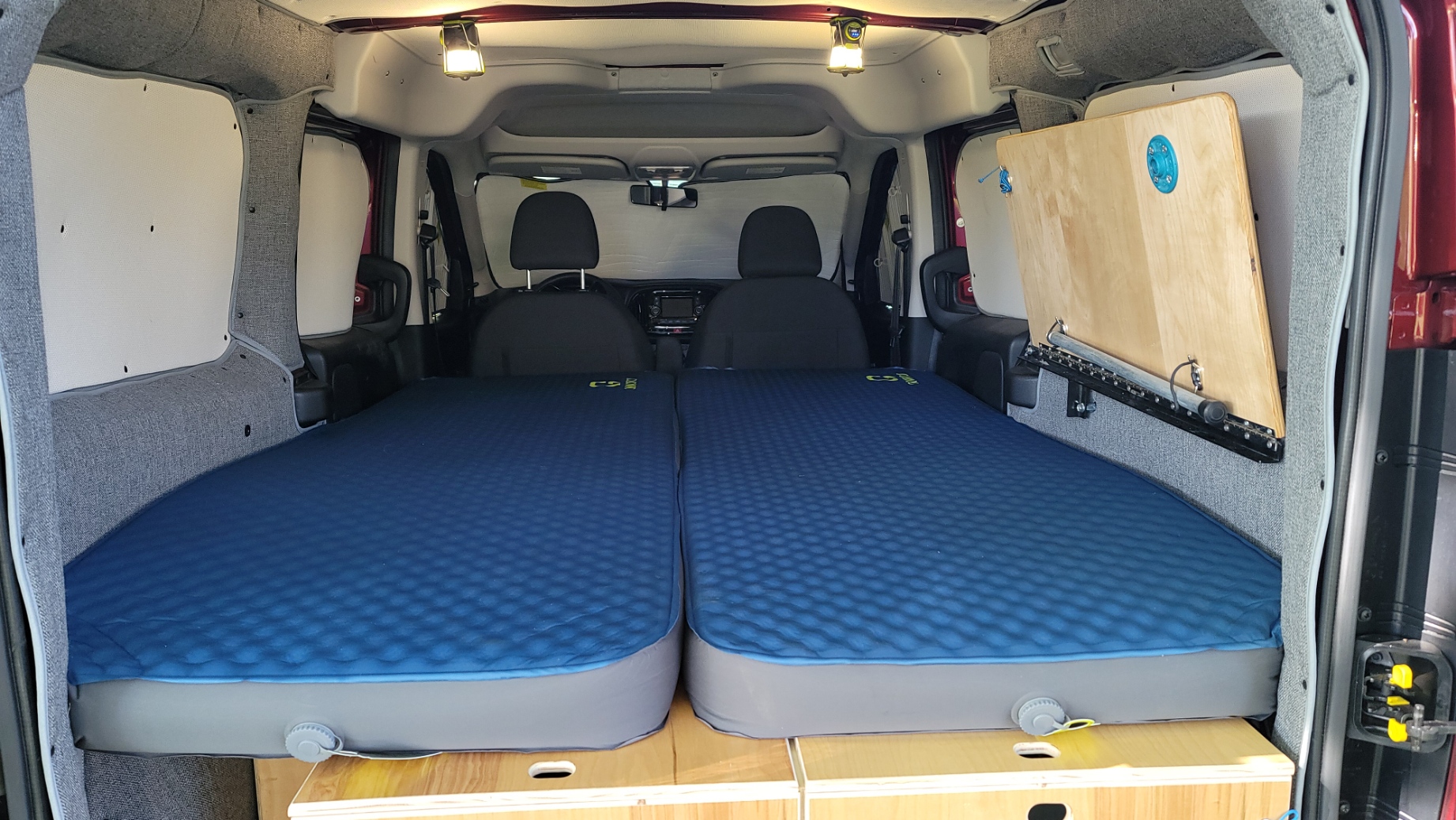 Inside an RV rental: a compact camper van with two blue sleeping pads creating a comfortable bed over wooden storage.. RAM Promaster CIty 2018