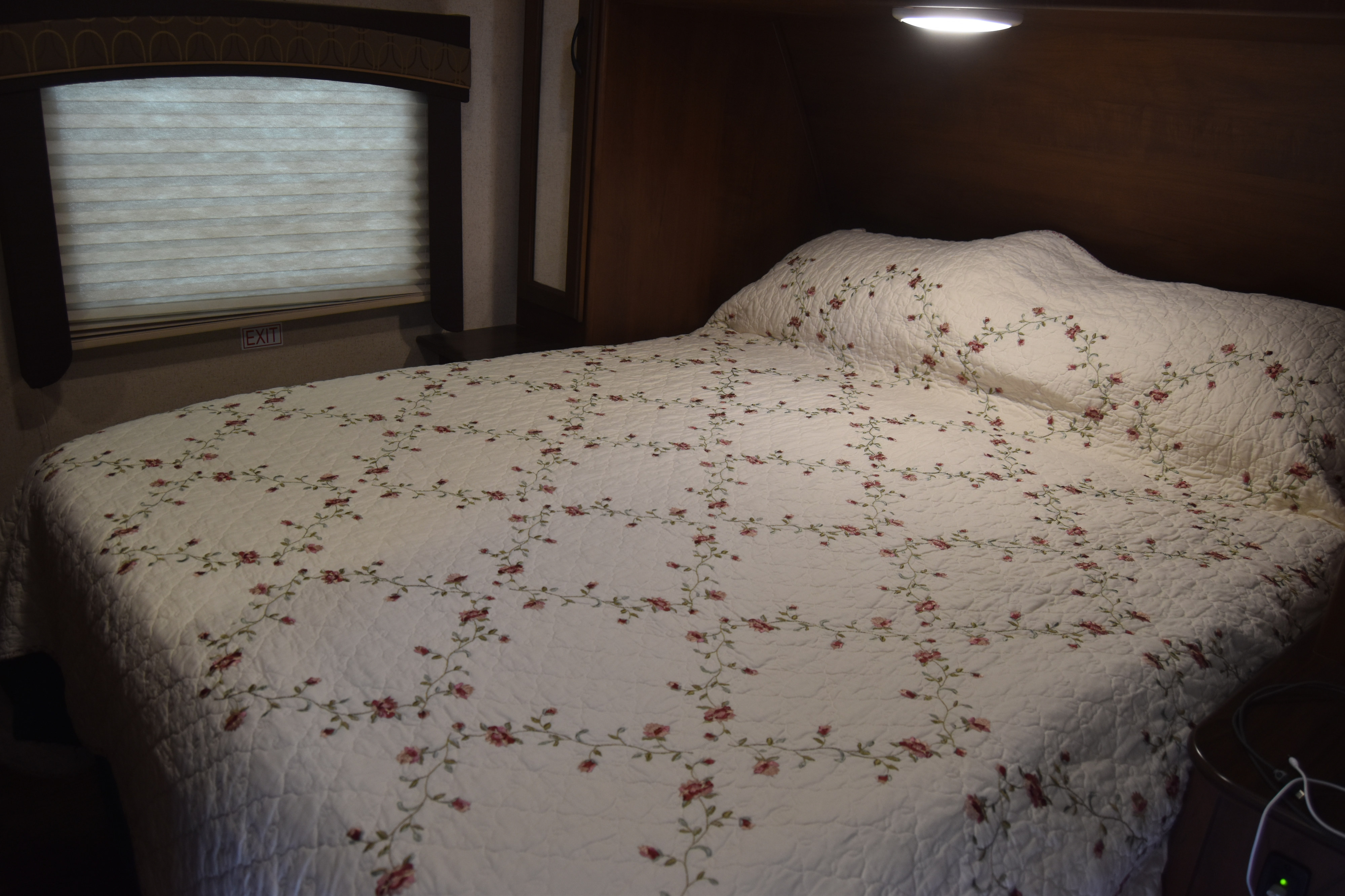 Inside an RV rental, a neatly made bed features a white quilt with a charming floral pattern. A window and exit sign are visible.. Jayco White Hawk Ultra Lite 2015