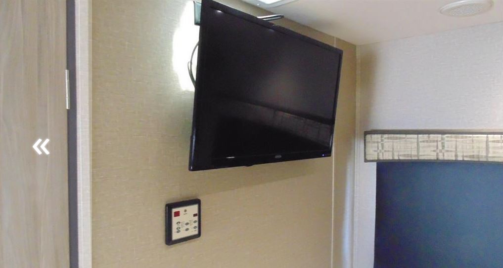 View of an RV rental interior with a wall-mounted TV, a control panel, and a window with a valance.. Dynamax Corp Isata 2016