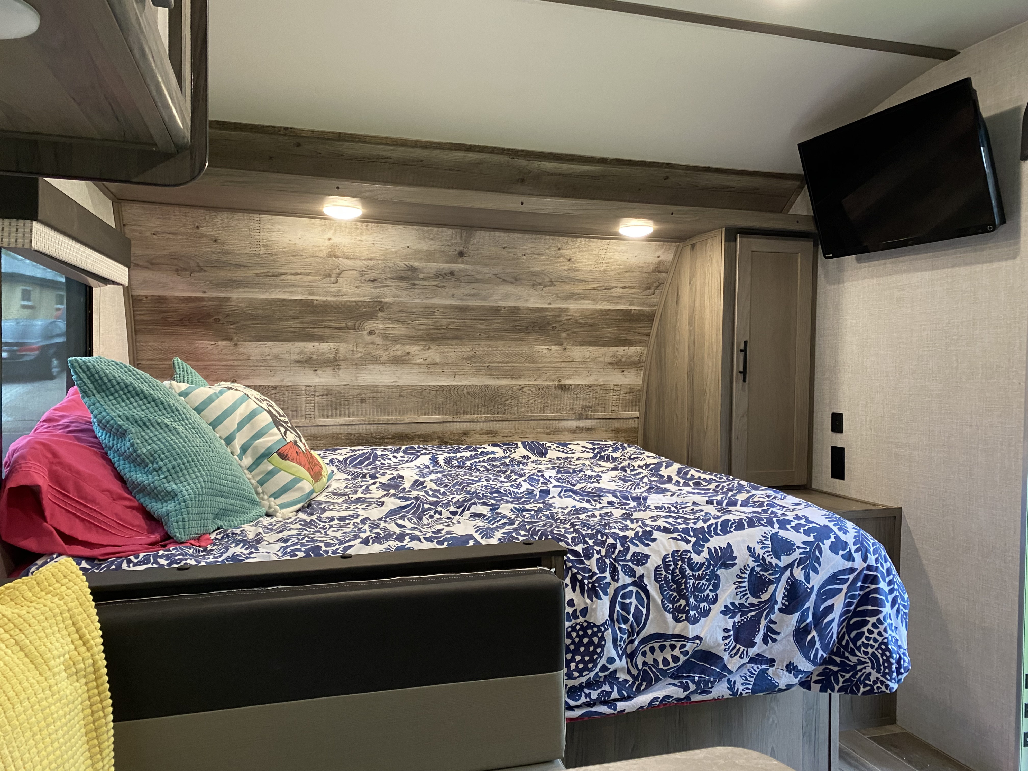 Cozy RV rental bedroom with a blue and white patterned bed, rustic wooden headboard, wall-mounted TV, and window.. Gulf Stream Amerilite 2021