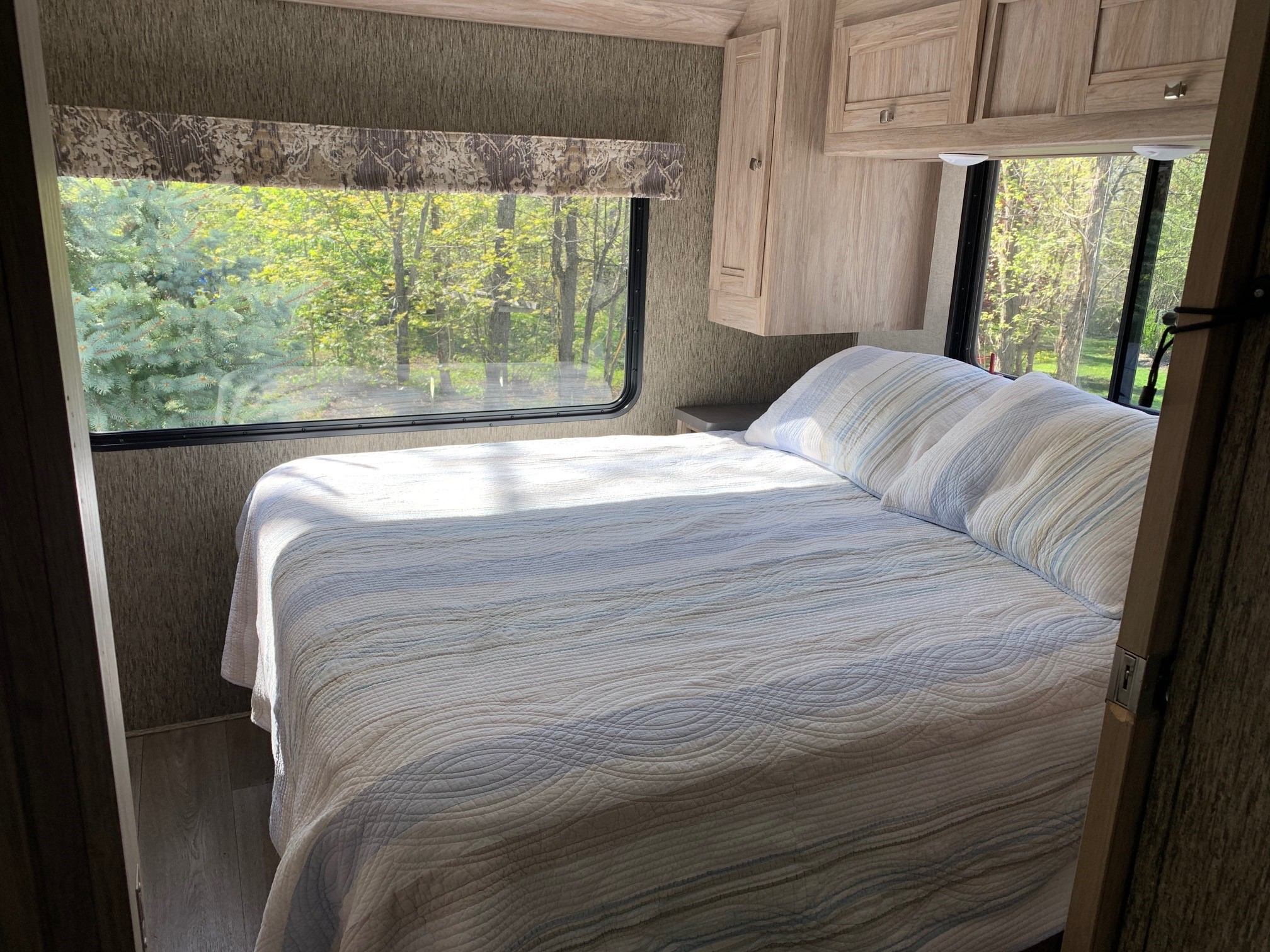 Cozy RV rental bedroom with sunlit bed, scenic windows overlooking green trees. Features overhead cabinets.. Forest River Sunseeker 2018