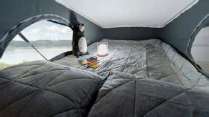 Inside a cozy RV rental pop-top, a comfy bed with grey quilted bedding and pillows faces a window with a serene lake view.. Winnebago Solis 2021