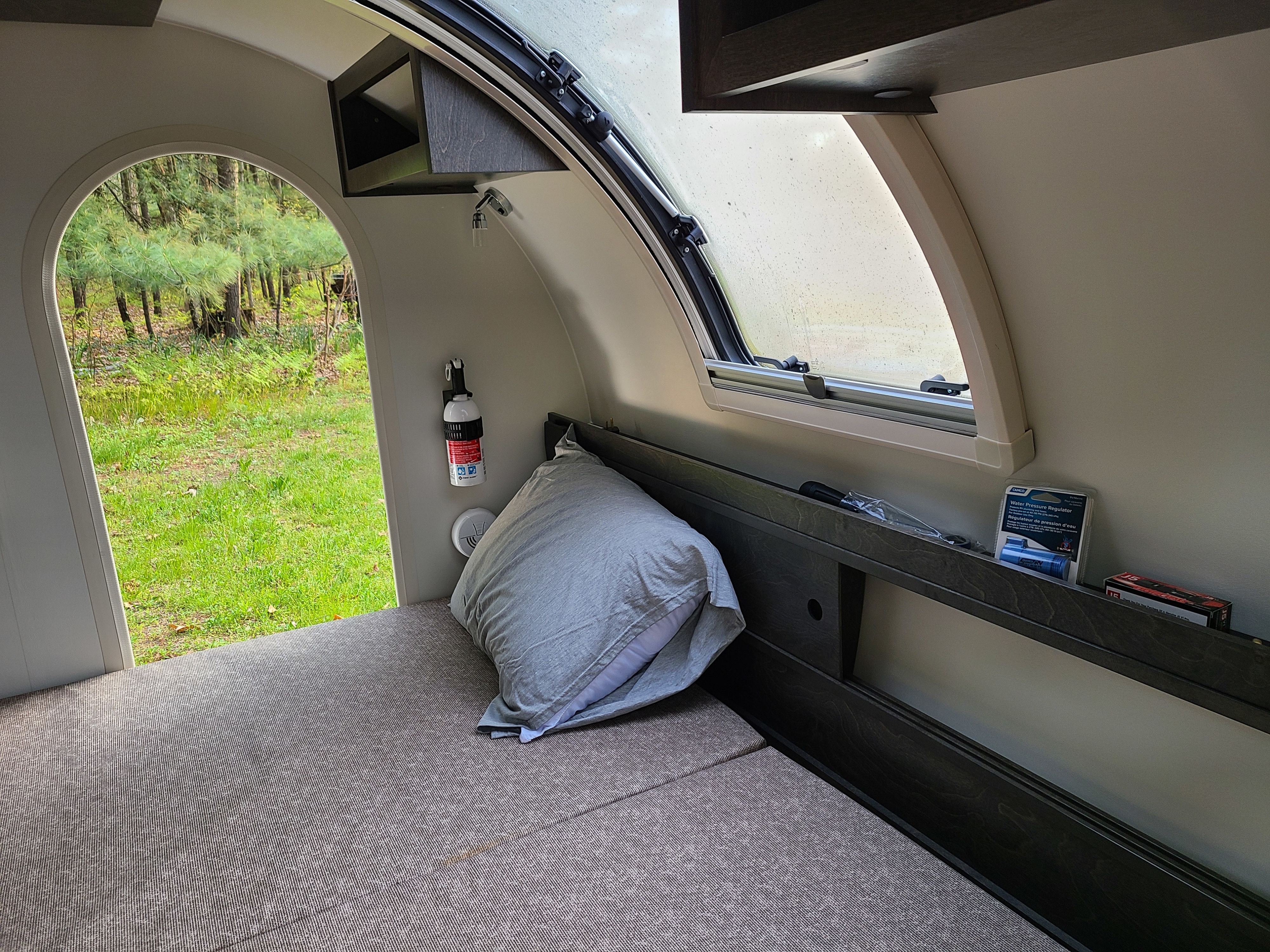 RV rental interior: bed, window, safety gear, and an open archway to a vibrant green forest view.. Nucamp Tag XL Boondock 2021