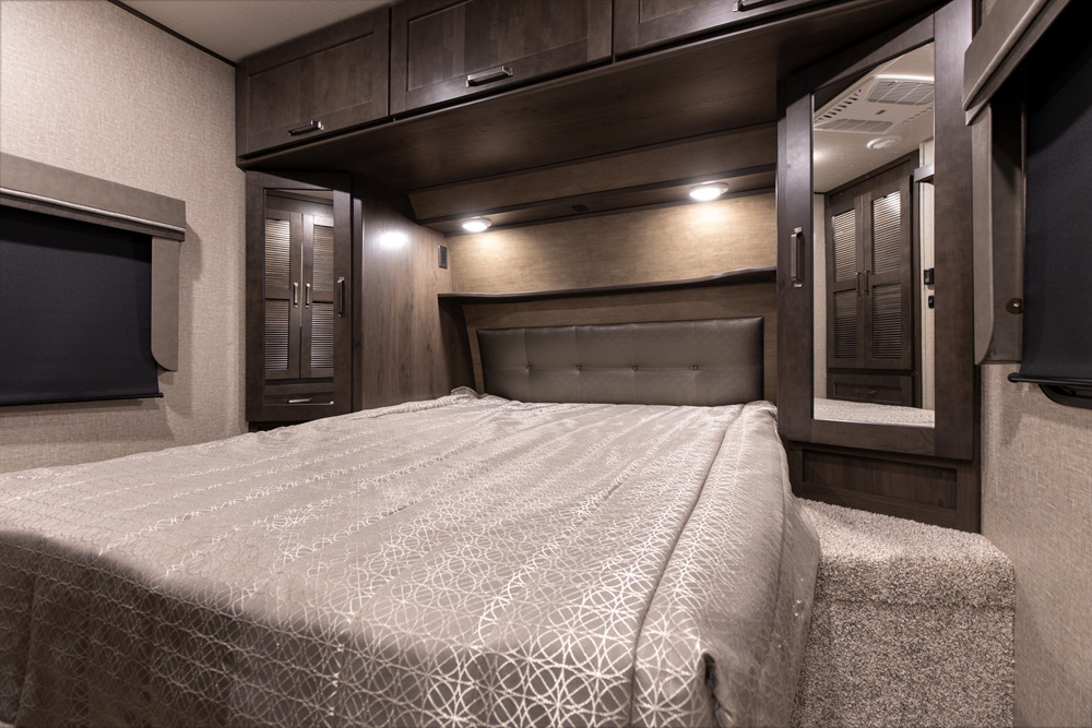 This spacious RV rental bedroom features dark wood cabinetry, a comfortable bed with patterned comforter, and a built-in mirror.. Grand Design Reflection 2020