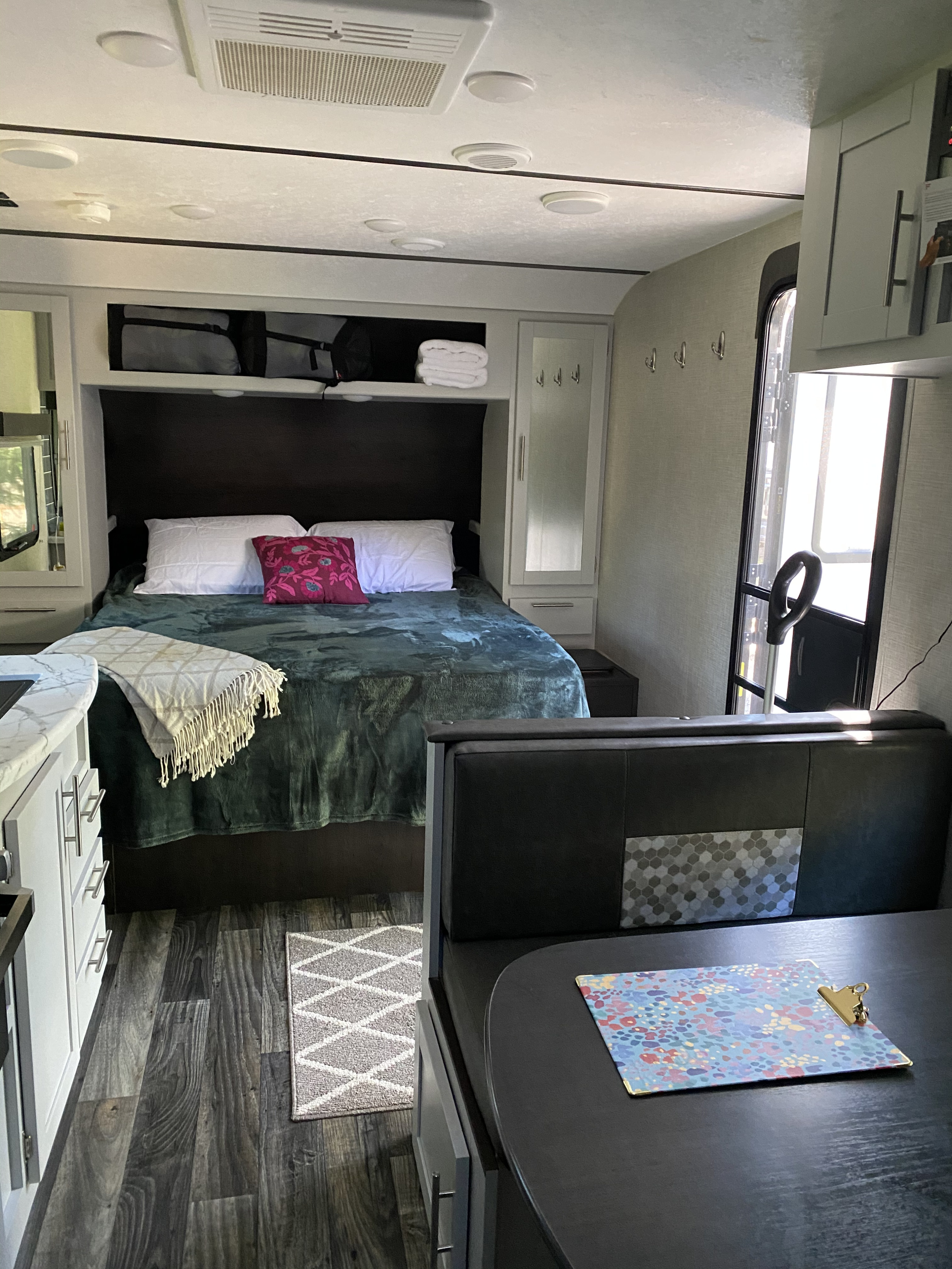 Bright RV rental interior showing a bed with a dark green blanket, a dinette booth, and a small kitchen counter.. Keystone Bullet 2020