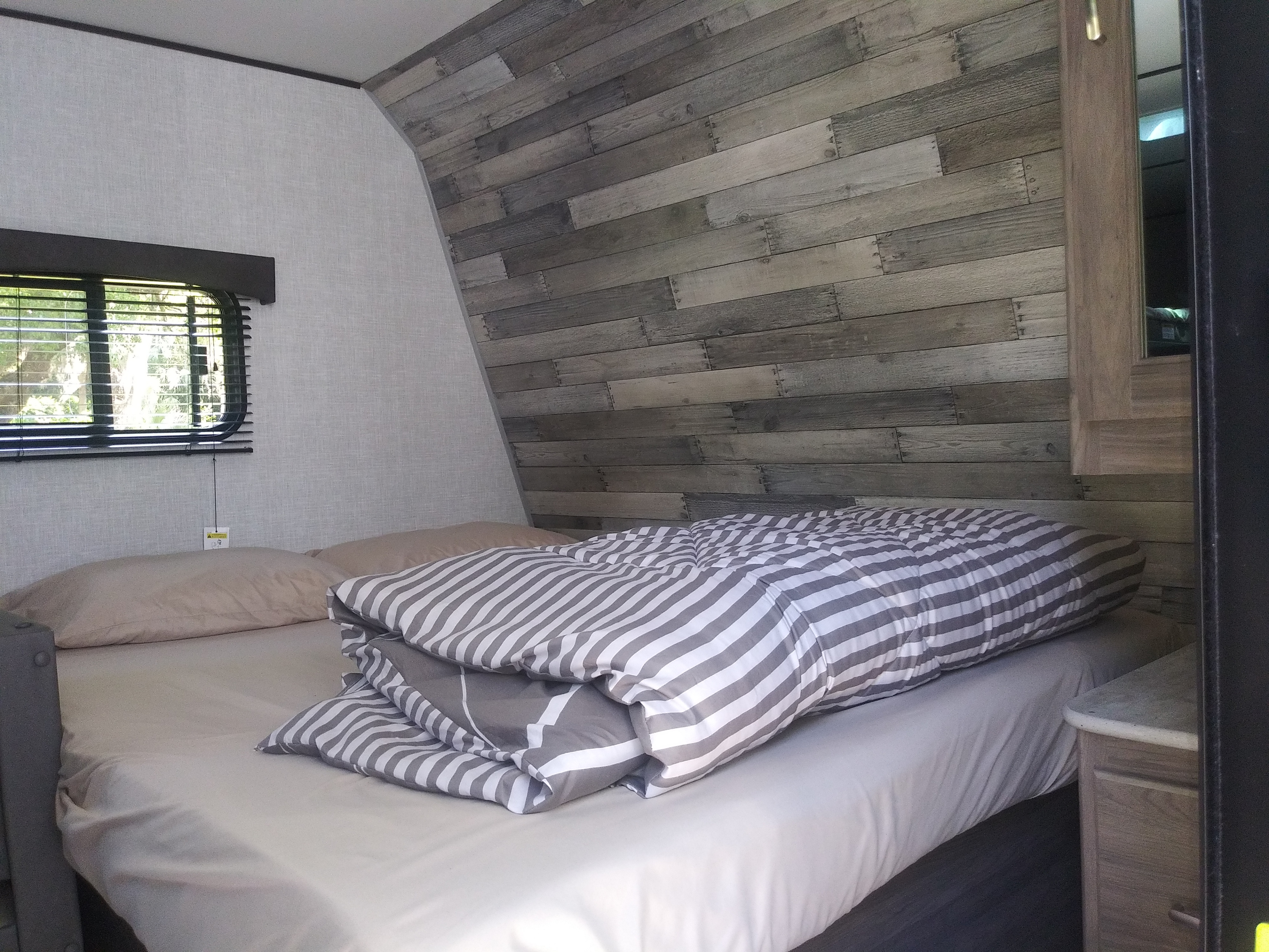 Inside an RV rental: a comfy bedroom with a grey wood accent wall, window blinds, and striped bedding. Ready for adventure.. Crossroads RV Zinger Lite 2021