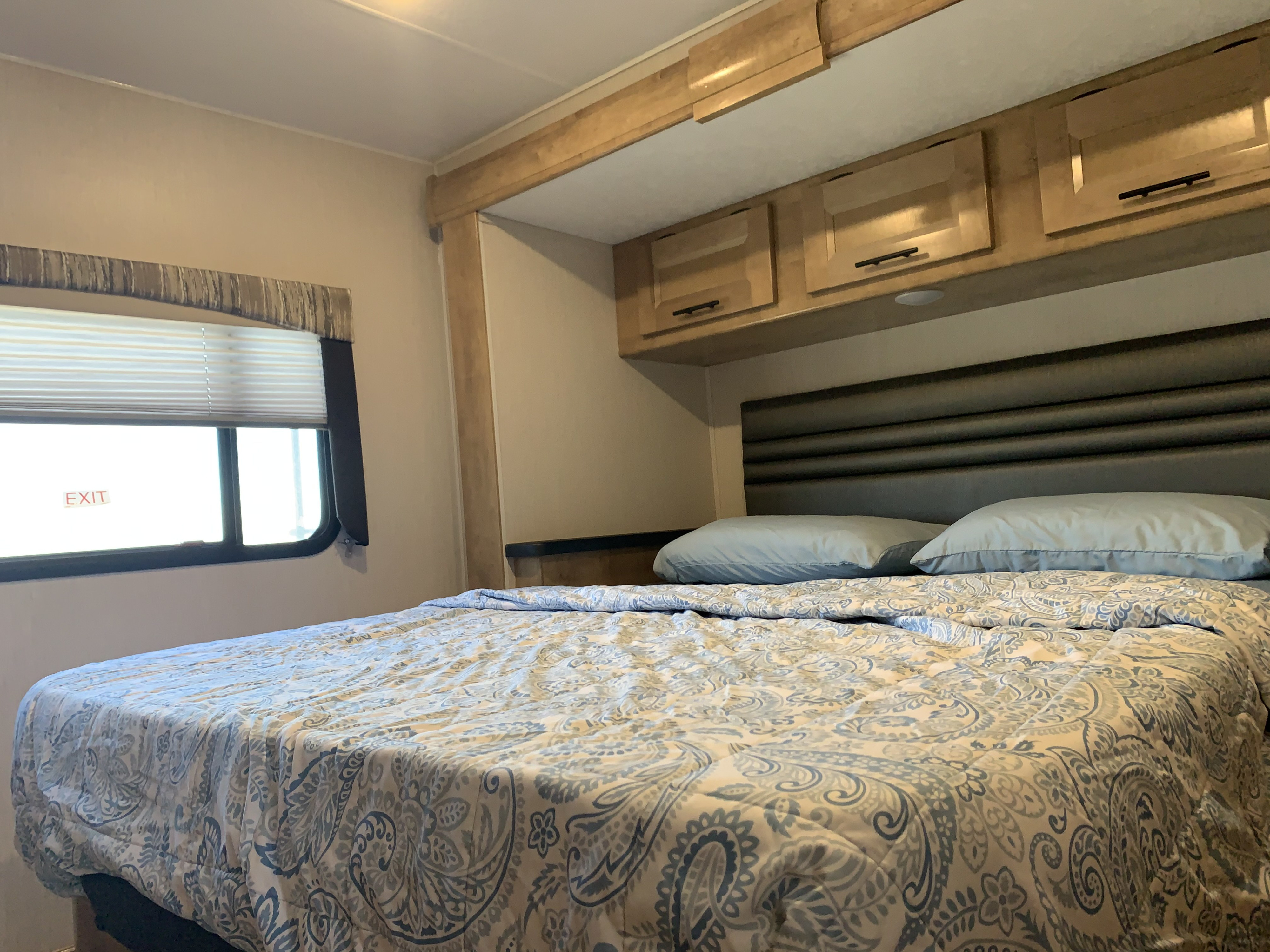 Cozy RV rental bedroom with a white & blue paisley bed, light wood cabinets, and an emergency exit window.. Coachmen Leprechaun 2020