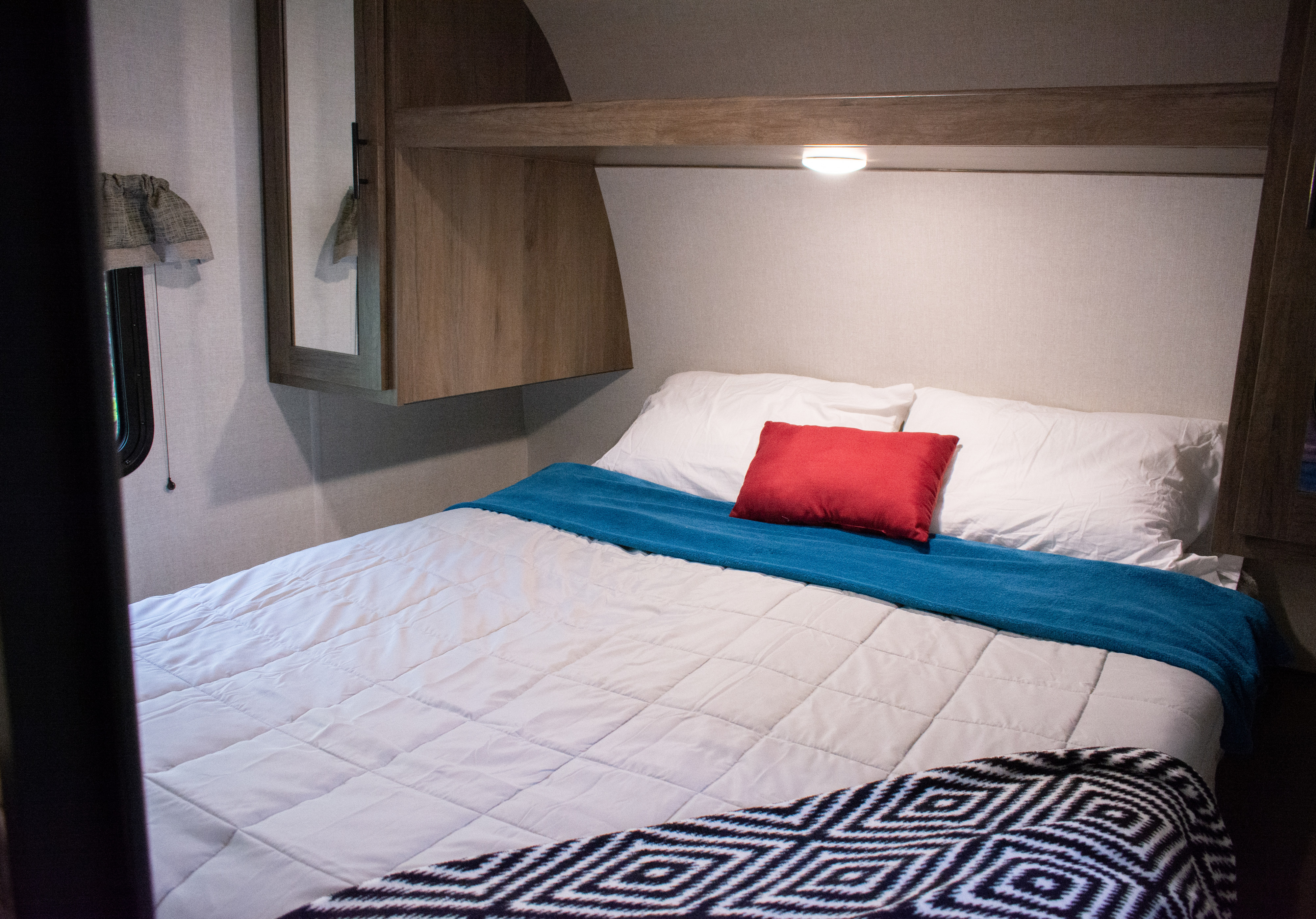 Modern RV rental bedroom: comfy bed with white linens, red pillow, blue throw. Built-in light & cabinetry.. Prime Time Avenger 2021