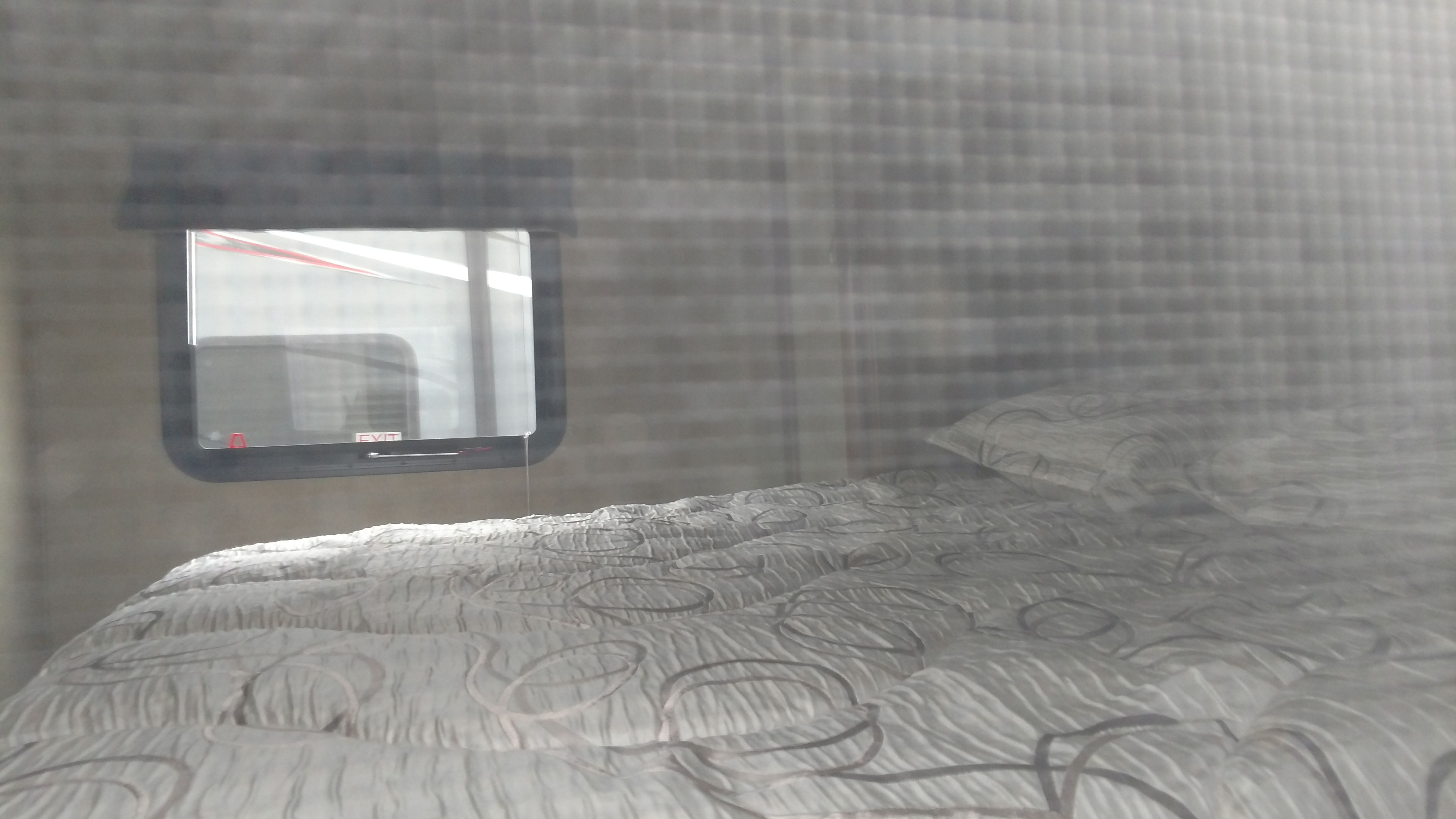 Inside an RV rental: a comfortable bed with a textured comforter and a window showing a reflected "EXIT" sign.. Forest River Vibe 2016