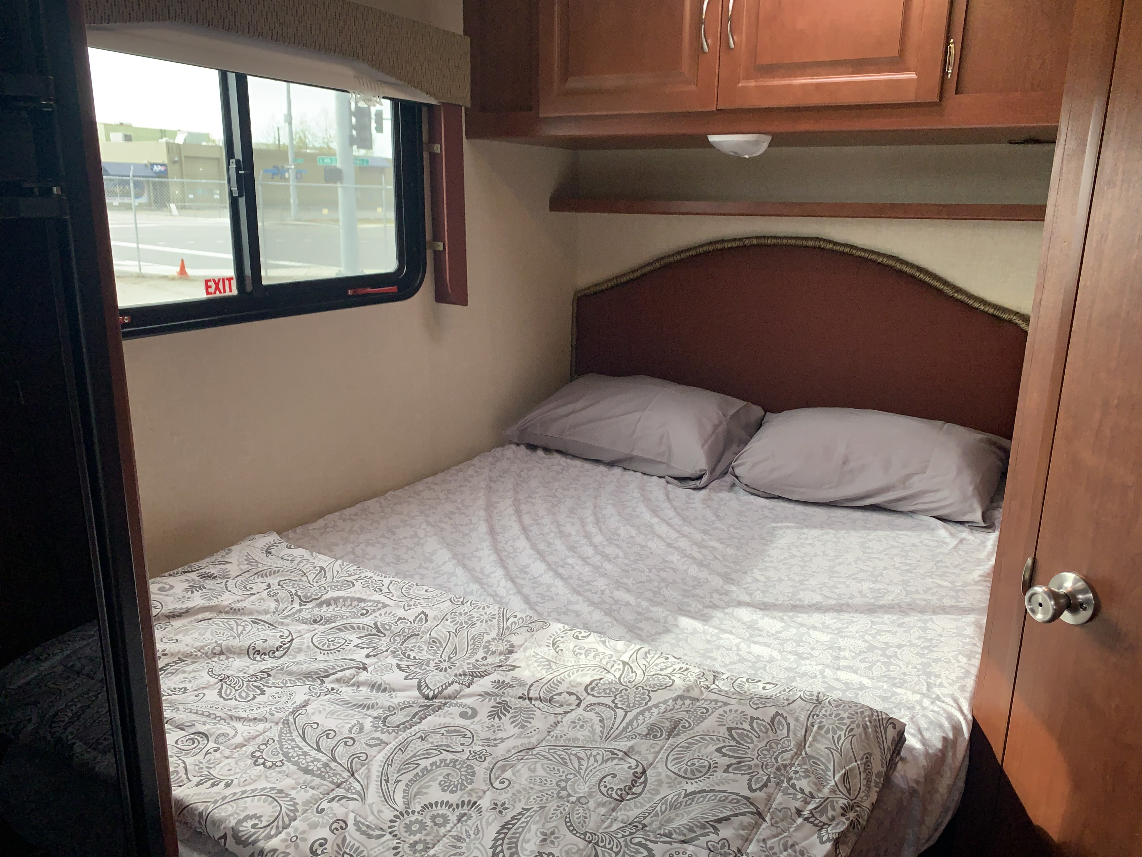 Cozy RV rental bedroom featuring a patterned bed with pillows, overhead cabinets, and a window view of the street outside.. Winnebago Minnie Winnie 2015