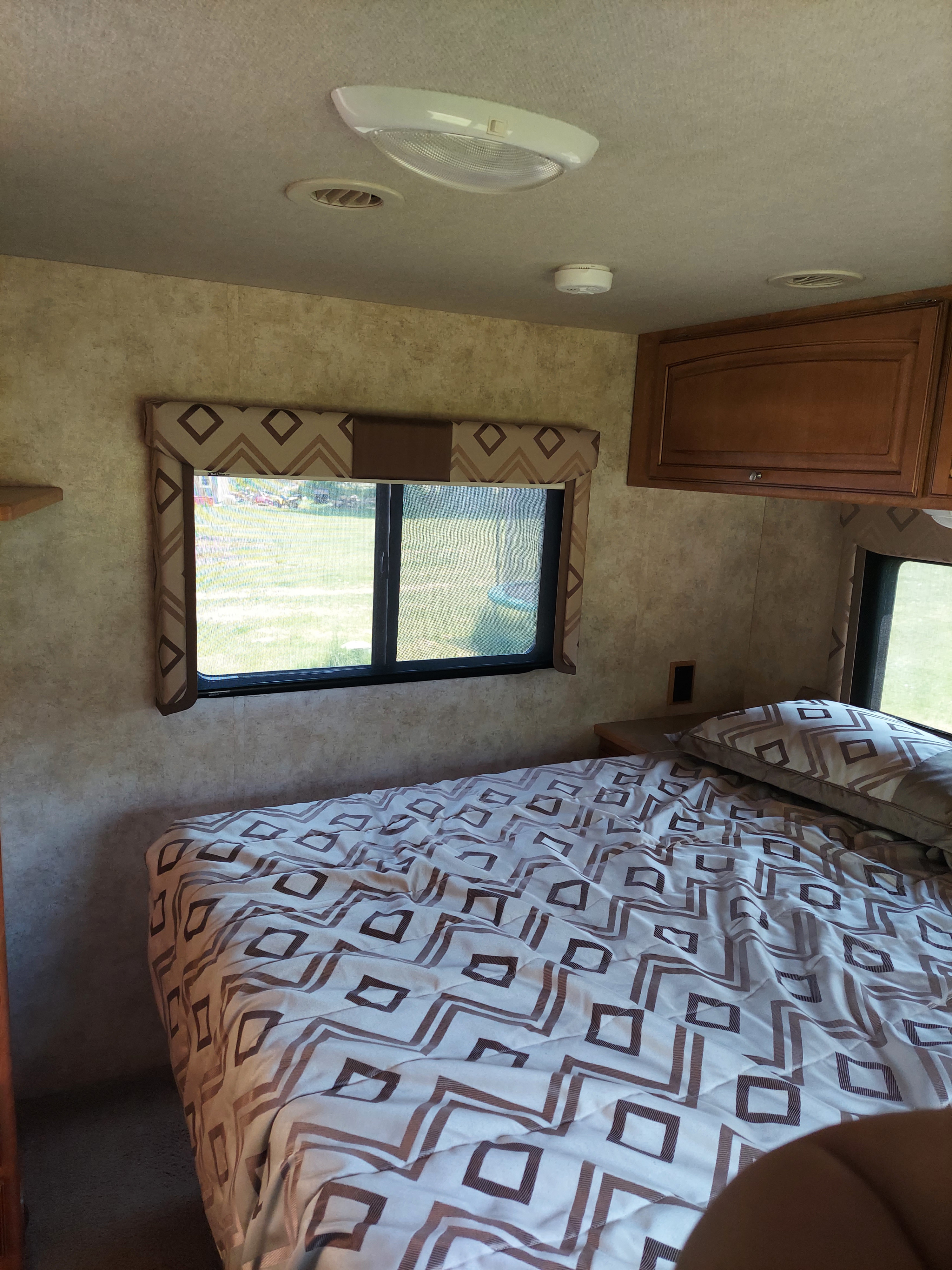 Cozy RV rental bedroom featuring patterned bedding, a window with a green view, and an overhead cabinet.. Winnebago Access 2011