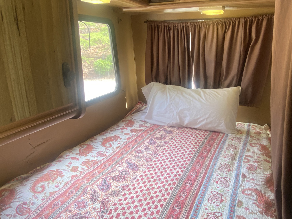 Cozy RV rental interior: bed with patterned quilt, white pillow, window, and brown curtains.. Fleetwood Pace Arrow 1984