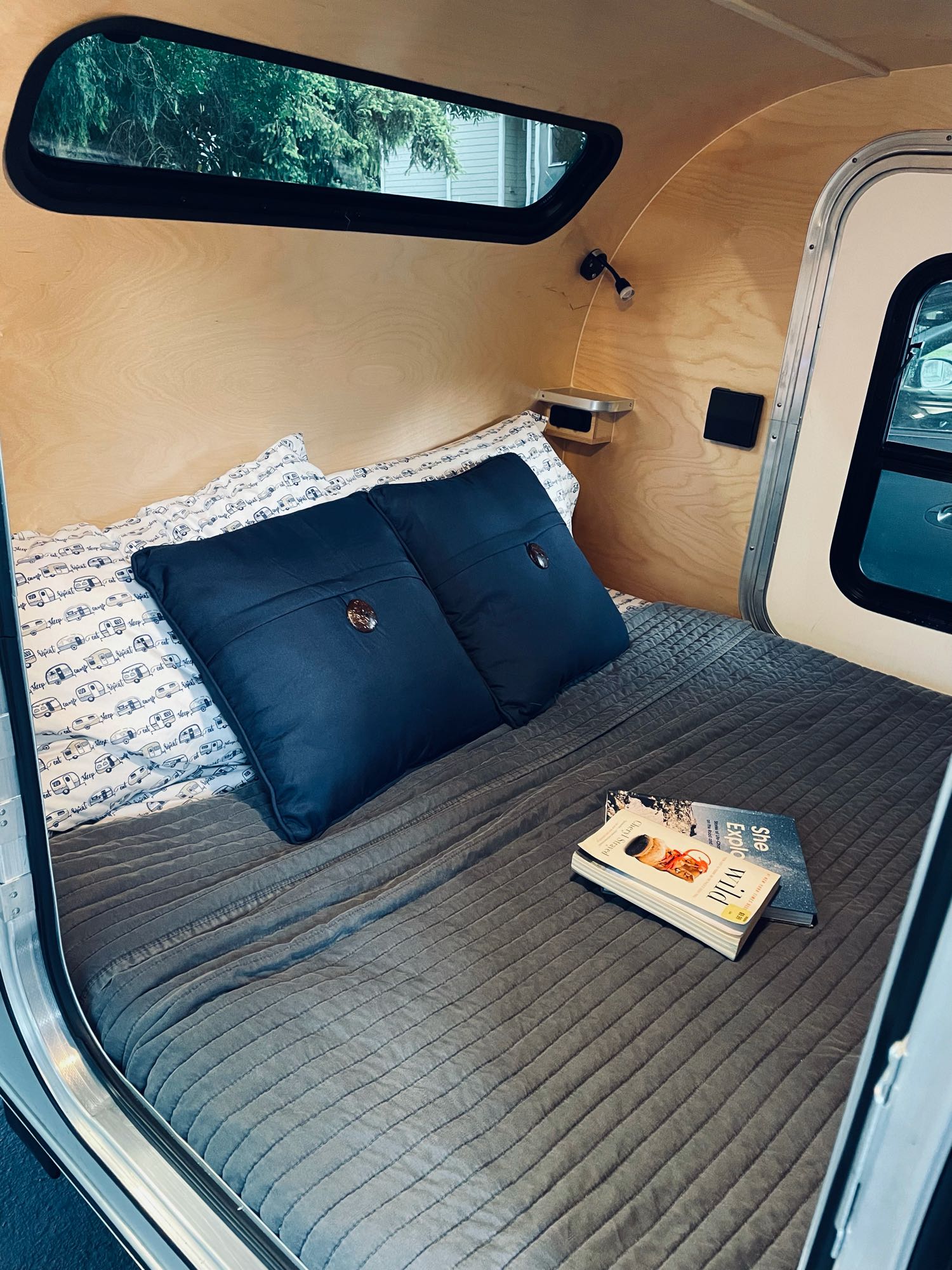 Cozy RV rental interior: a bed with gray quilt, blue pillows, and books like 'Wild' on light wood walls.. Aero Teardrops Teardrop Camper 2022