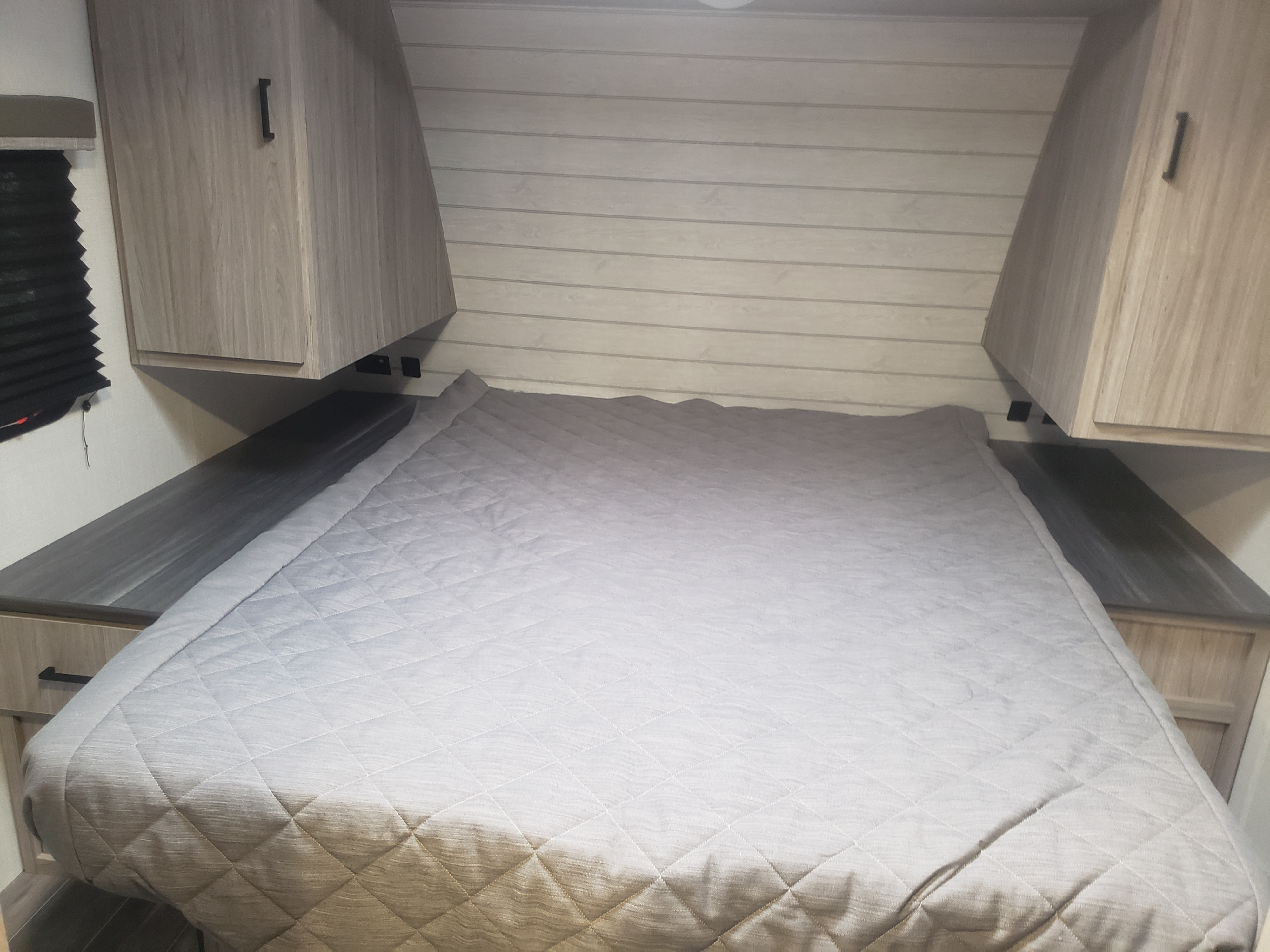 An RV rental bedroom, neatly arranged, with a light grey quilted bed, built-in side tables, and overhead storage cabinets.. Keystone Passport 2021