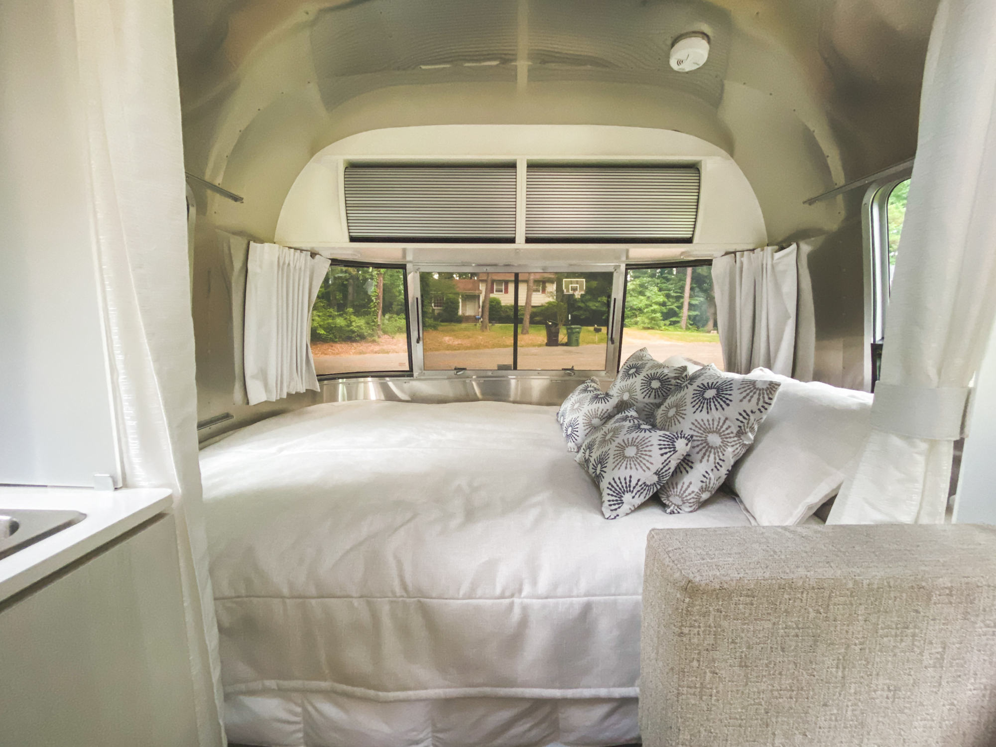 Explore this Airstream RV rental's bedroom: a plush white bed, decorative pillows, and expansive rear windows overlooking a residential street.. Airstream Other 2021