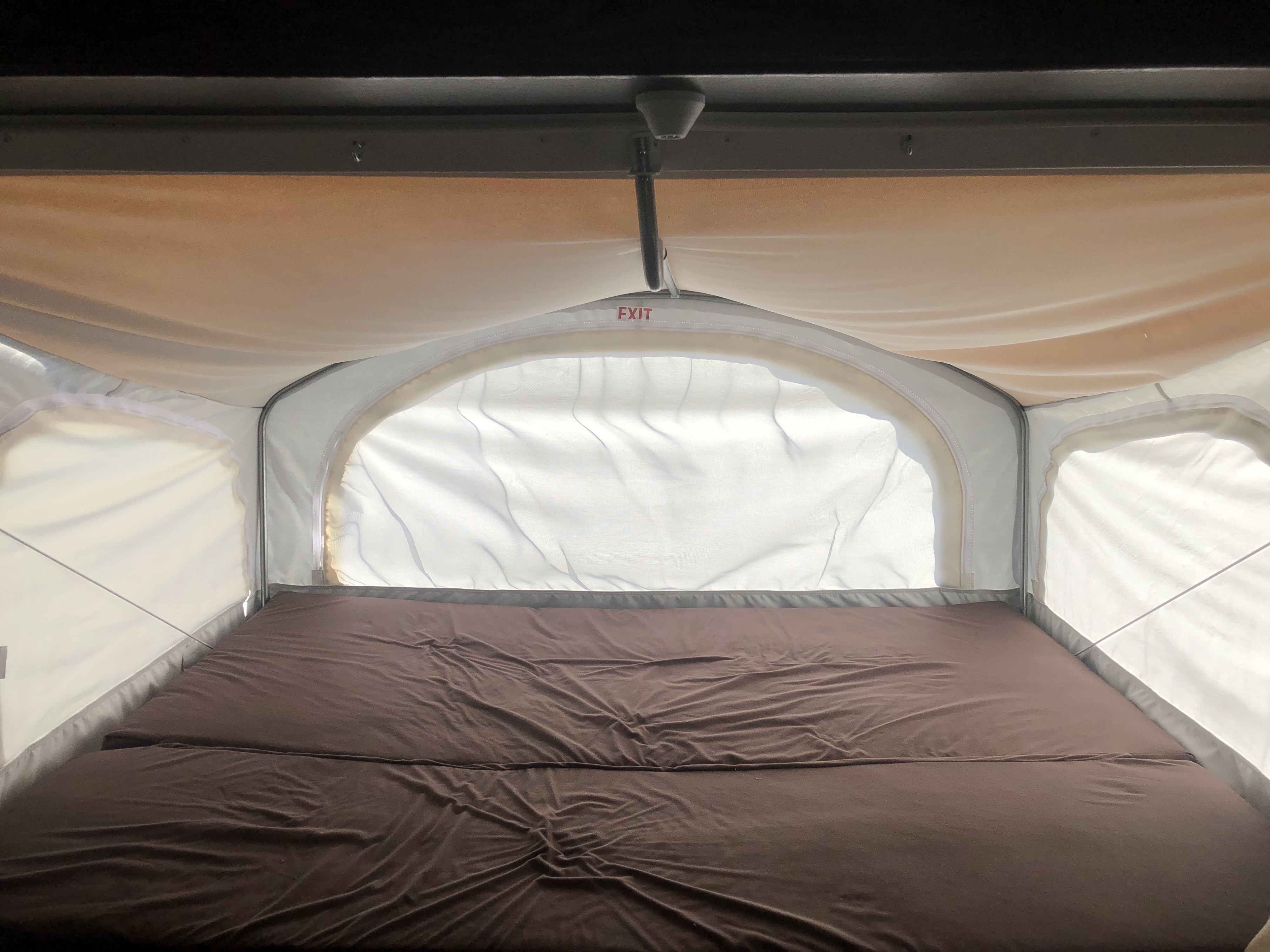 Inside an RV rental: Brown mattress fills this pop-up camper's cozy sleeping area, with fabric walls & exit sign.. Dutchmen Kodiak 2019