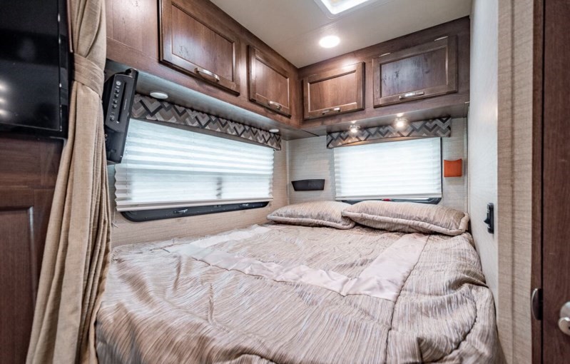 RV rental interior: a cozy bedroom with a comfortable bed, dark wood cabinets, windows with blinds, and a mounted TV.. Jayco Redhawk 2018