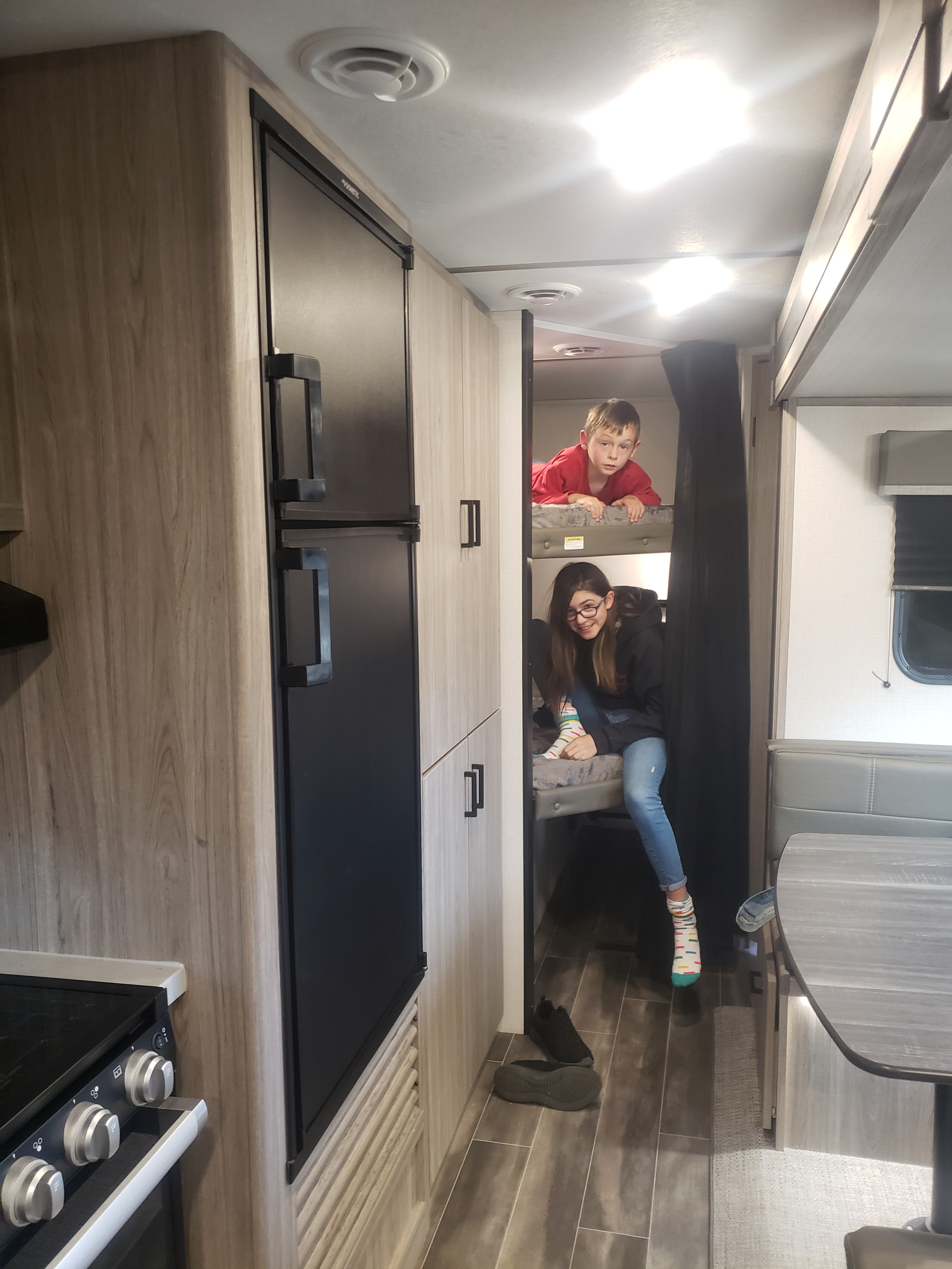 Inside an RV rental, two kids enjoy the bunks. A kitchen and dining area are visible, showcasing the compact, family-friendly space.. Keystone Passport 2021